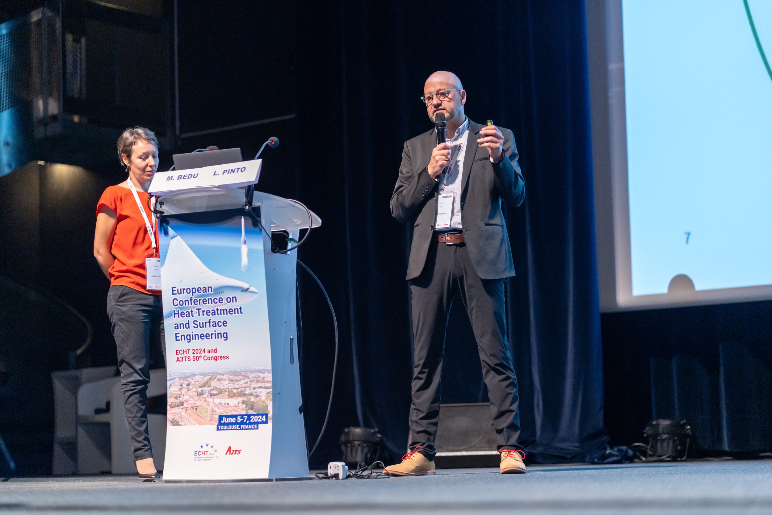 Conférence en cours avec deux intervenants sur scène, un homme et une femme. L'homme tient un micro. Un podium affiche le texte "European Conference on Heat Treatment and Surface Engineering."
