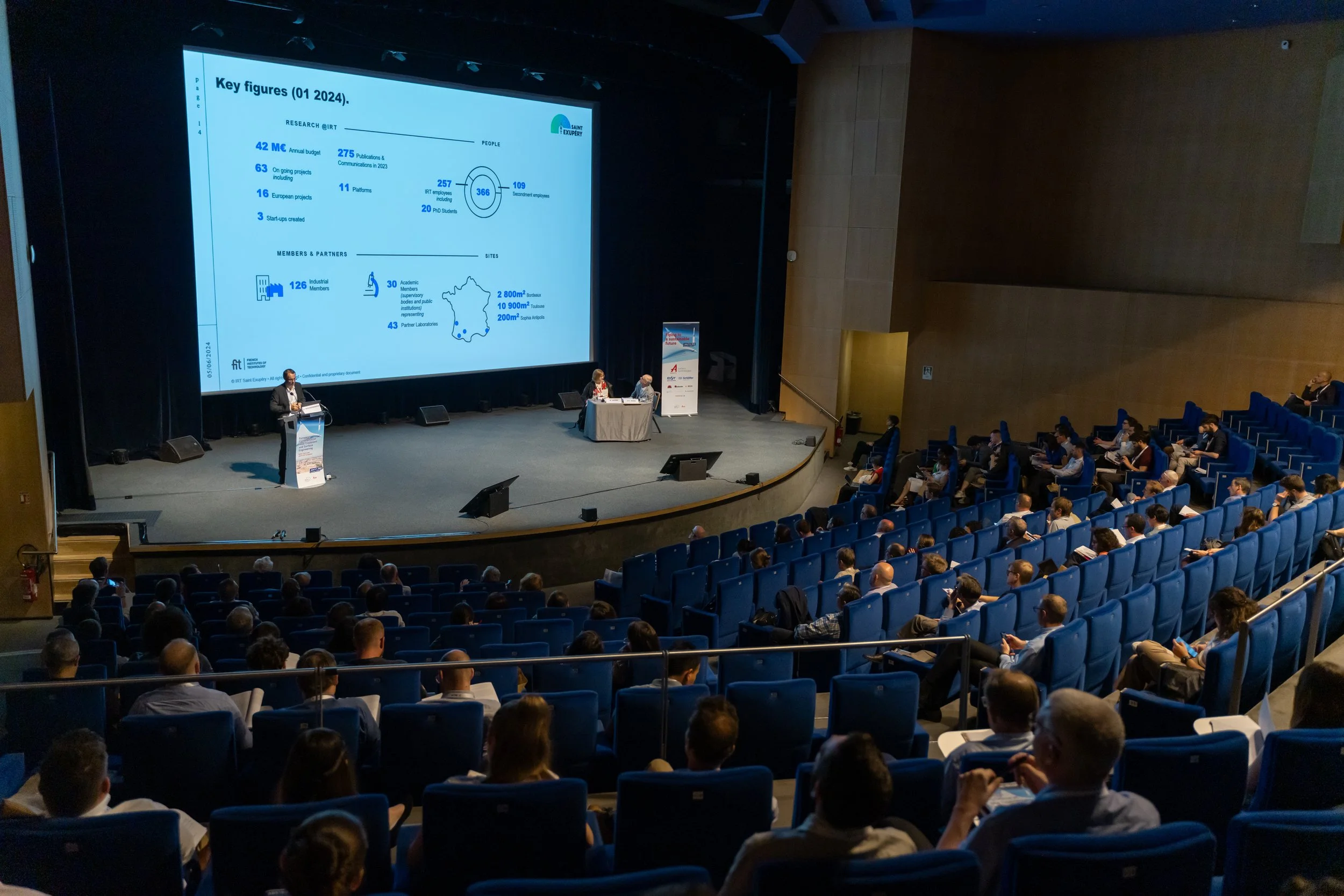 Une conférence dans un amphithéâtre avec un présentateur sur scène et un grand écran affichant des données clés de recherche. Le public est assis dans des sièges bleus, écoutant attentivement.