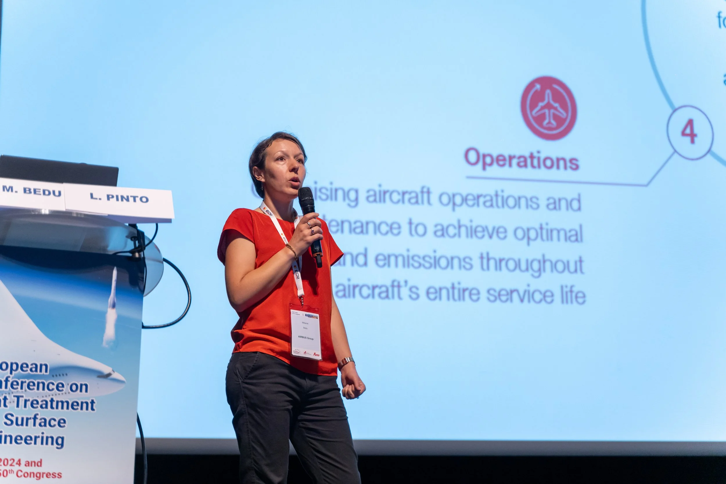 Une femme en chemise rouge parle dans un micro lors d'une conférence, debout près d'un pupitre. À l'arrière-plan, une présentation projetée sur un écran mentionne "Operations" et des détails sur l'optimisation des opérations aériennes.
