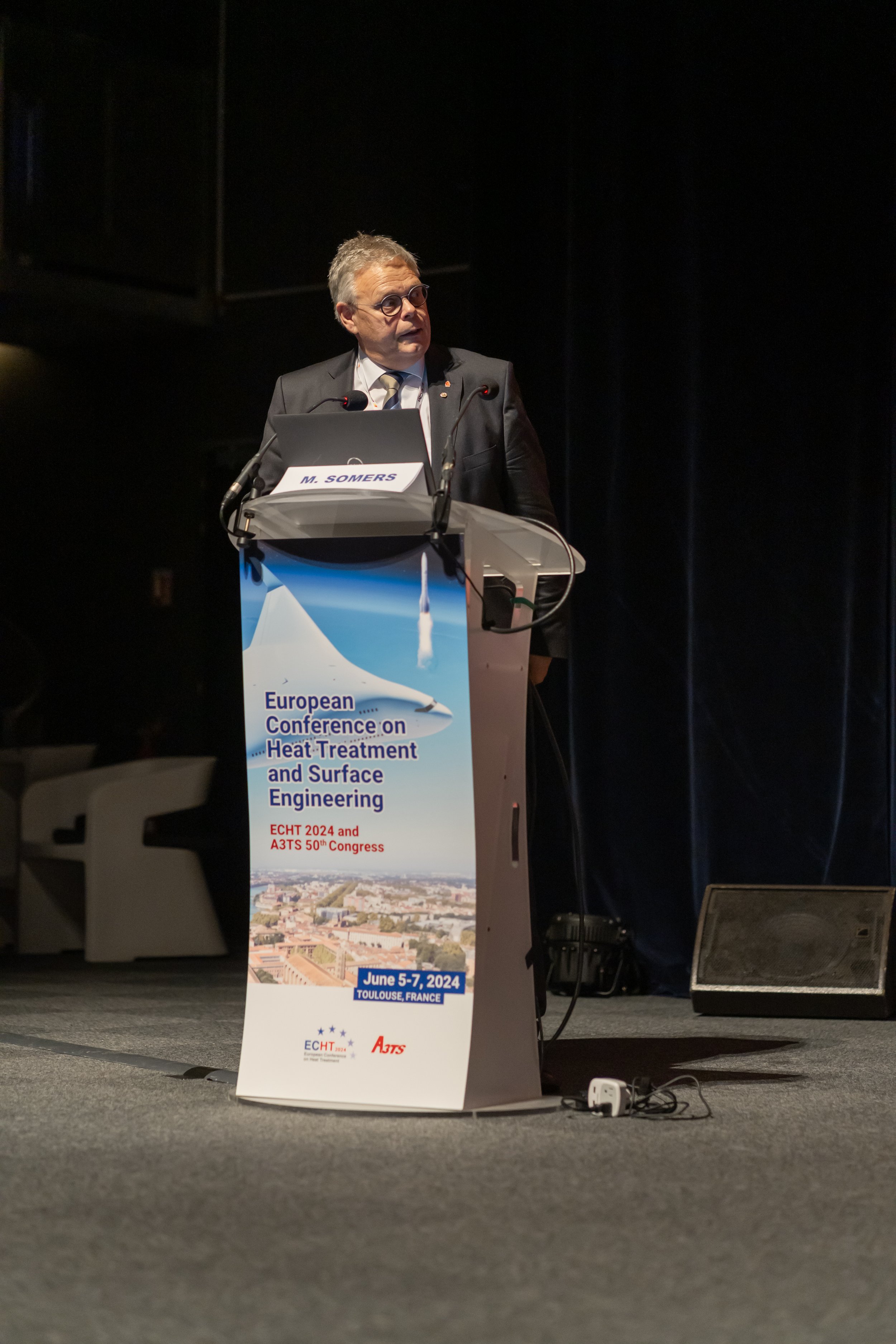 Un homme en costume présente à une conférence sur le traitement thermique et l'ingénierie de surface en Europe. Le podium affiche les détails de l'événement "European Conference on Heat-Treatment and Surface Engineering" qui se tiendra du 5 au 7 juin