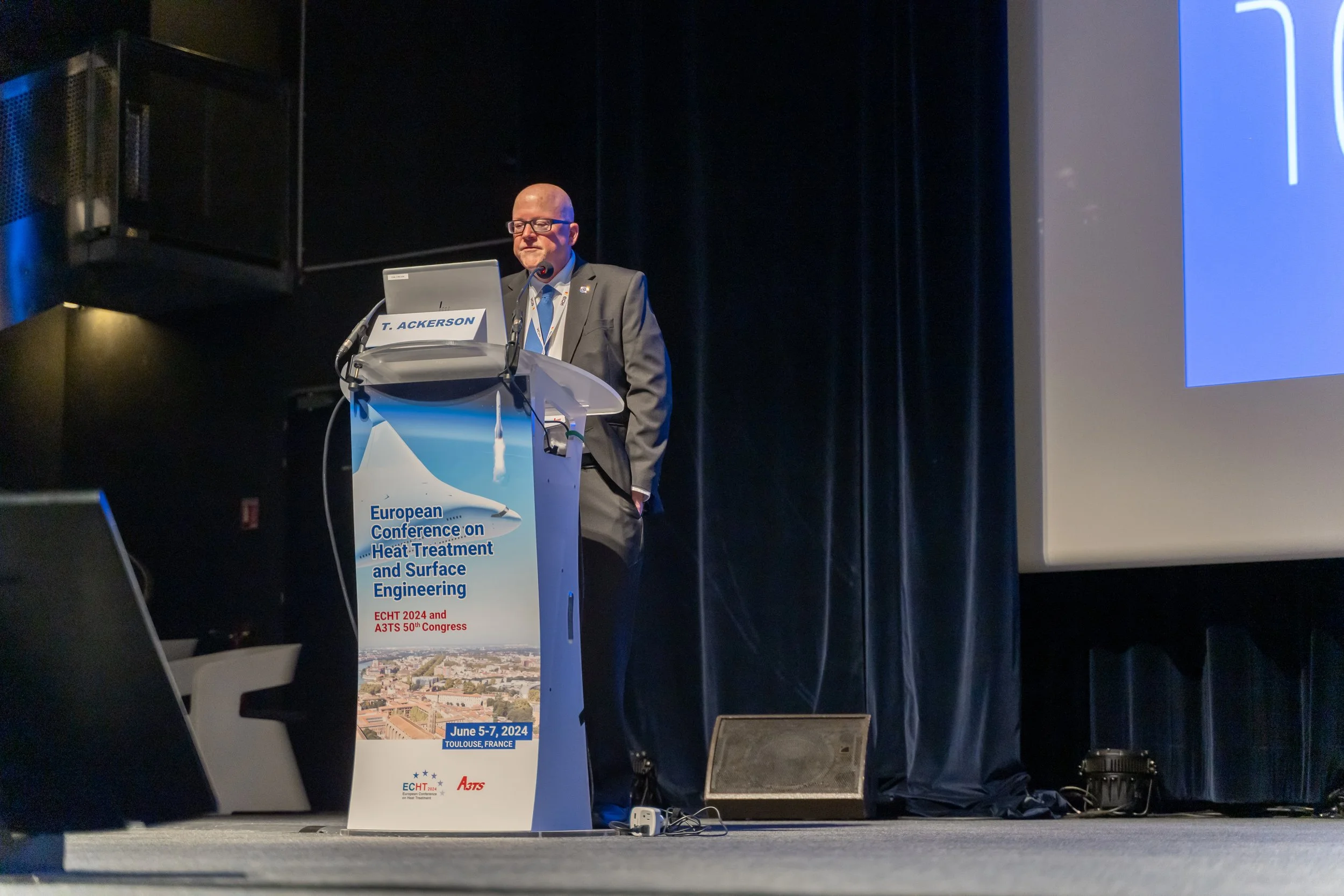 Homme en costume debout à un podium lors d'une conférence intitulée "European Conference on Heat-Treatment and Surface Engineering" à Toulouse, France, prévue du 5 au 7 juin 2024.