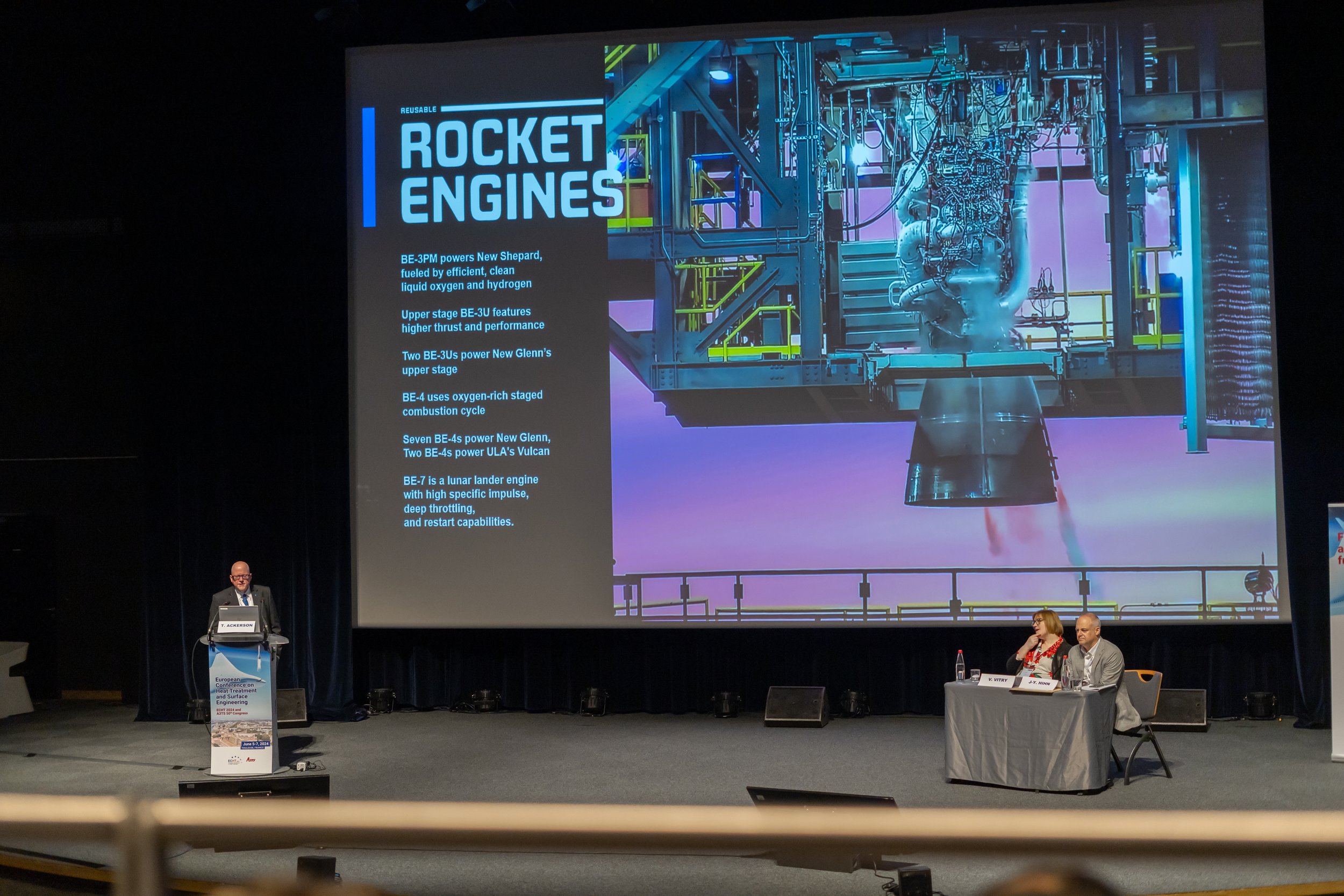 Conférence présentant des moteurs de fusées avec une diapositive affichant des détails techniques sur l'écran. Un intervenant se tient derrière un pupitre et deux personnes sont assises à une table sur l'estrade.
