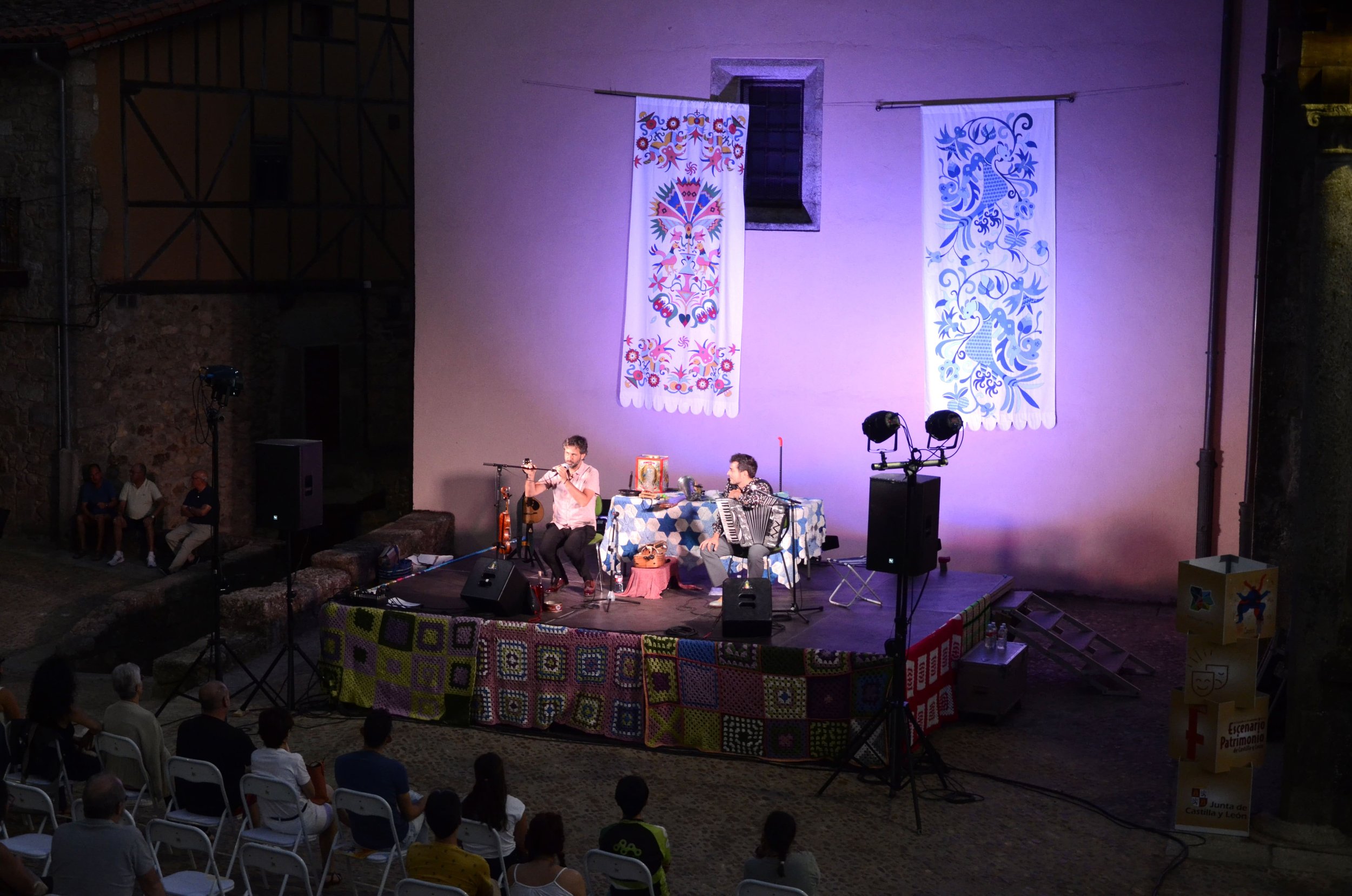 Espectáculo de música en un escenario al aire libre con decoraciones coloridas. Dos músicos tocan instrumentos tradicionales, rodeados de música y pintura en un entorno histórico.