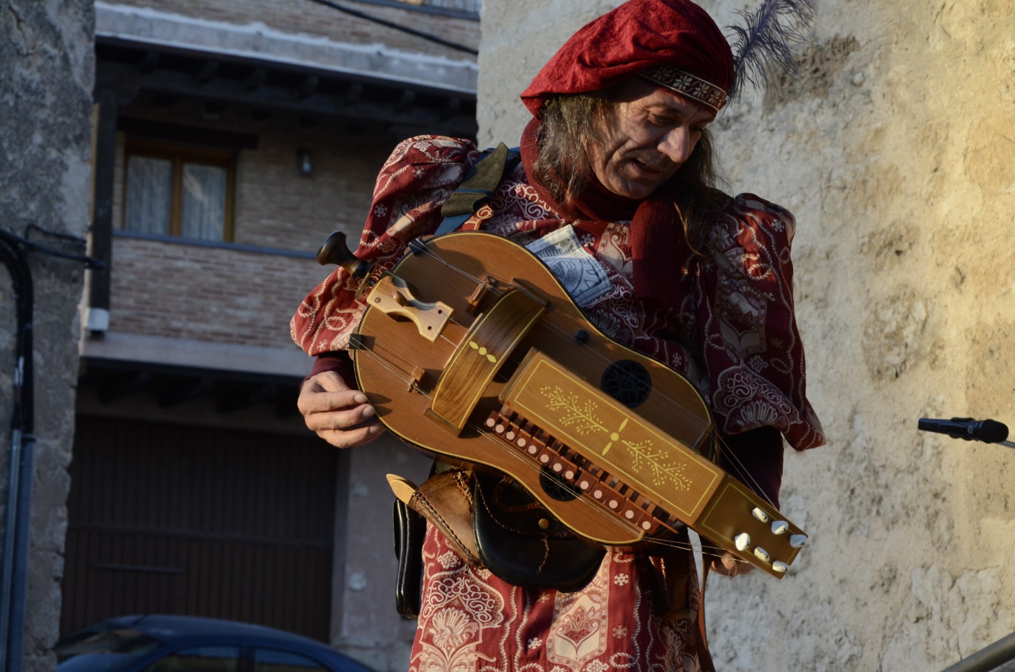 Un hombre vestido con ropa de estilo medieval o renacentista tocando un instrumento musical, posiblemente una rabel, en un entorno urbano con paredes de piedra y edificios antiguos.