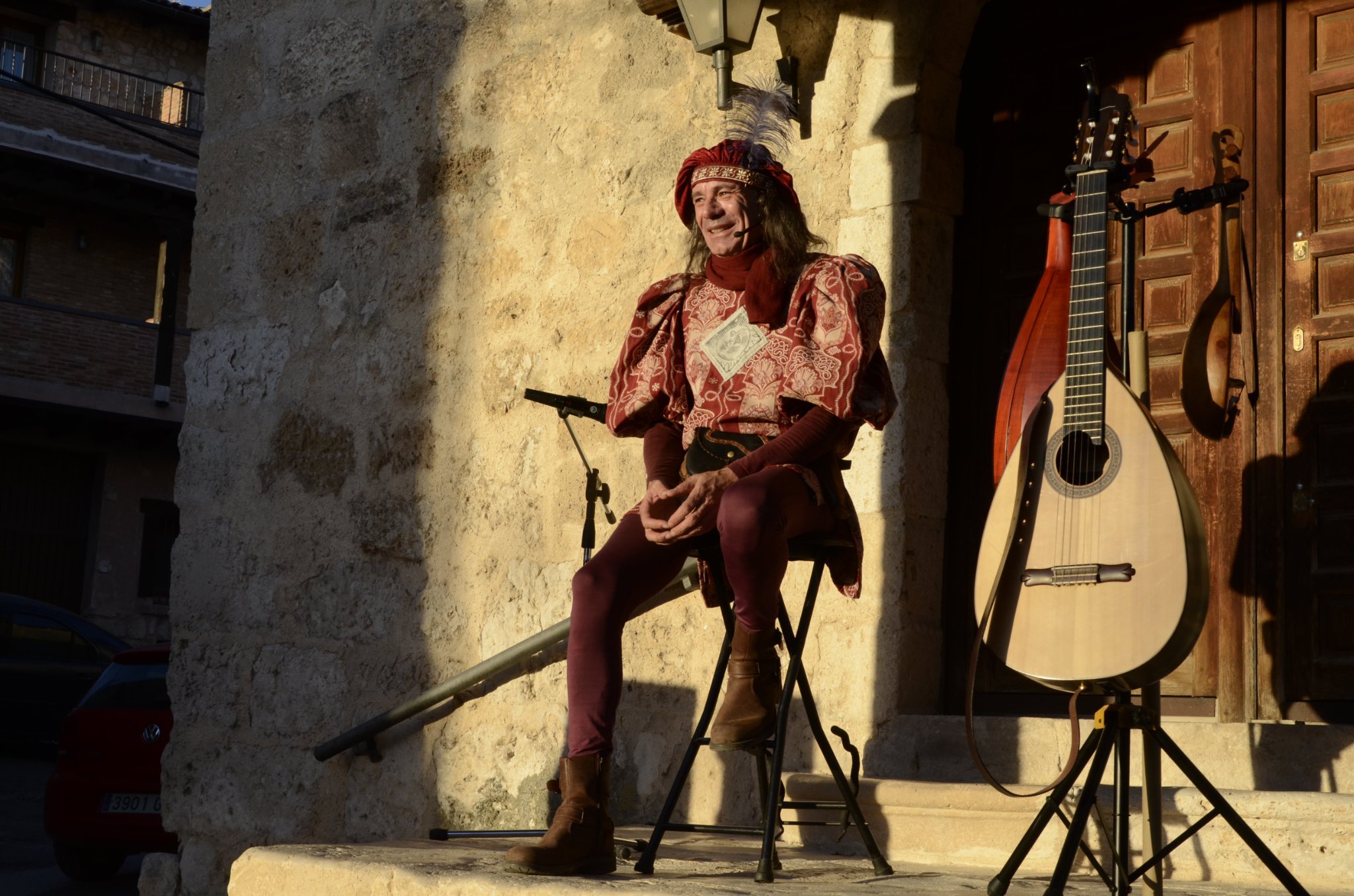 Persona disfrazada de caballero medieval sentado en una silla frente a un fondo de piedra, con una guitarra acústica y una segunda guitarra colgada a la derecha, en un escenario al aire libre.
