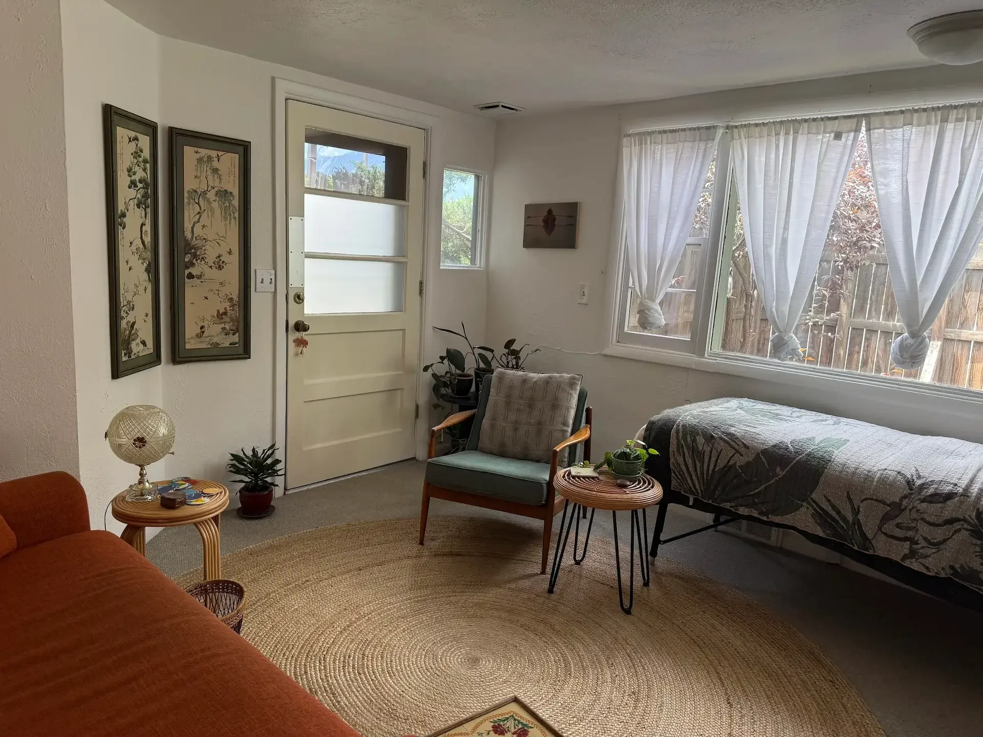 A cozy therapy office with a window and white curtains, a small table with a potted plant, an armchair, a treatment table with a blanket, and a framed painting on the wall.