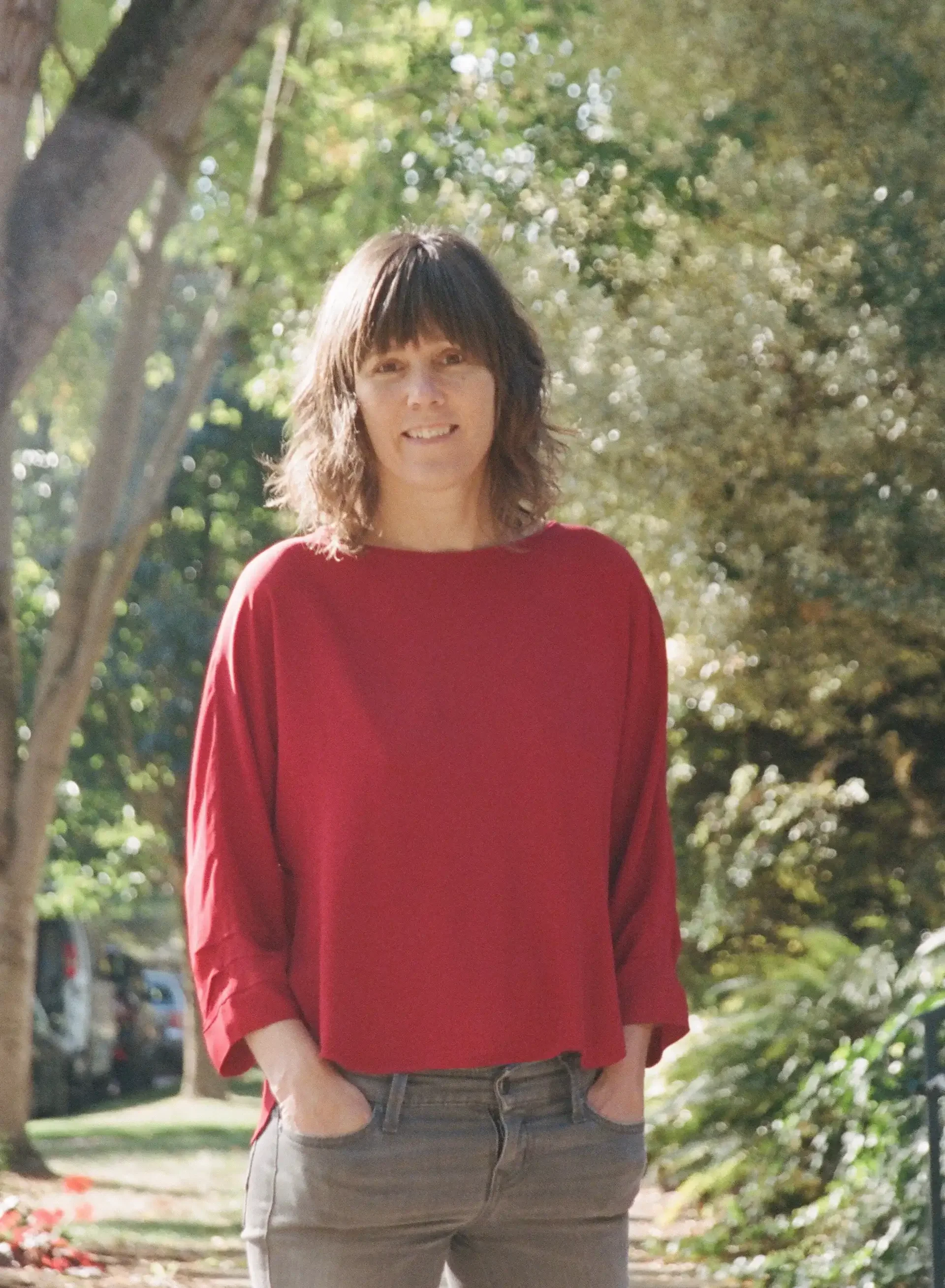 A woman wearing a red long-sleeve shirt paired with gray pants, standing outdoors on a sunny day with trees and greenery in the background.