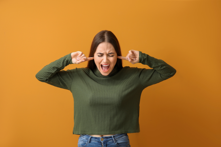 Woman standing, plugging her ears, and a pained expression on her face.