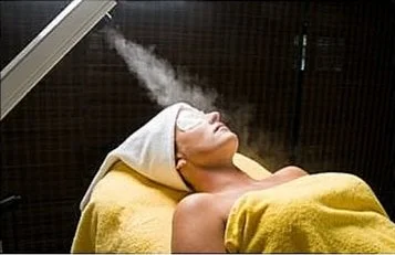 Woman lying on a spa bed with a towel on her head and pads on her eyes. A machine is blowing steam on her face to open up her pores.