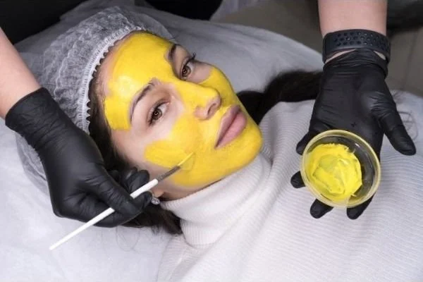 A woman lying on down, getting a chemical peel solution applied to her face with a brush.