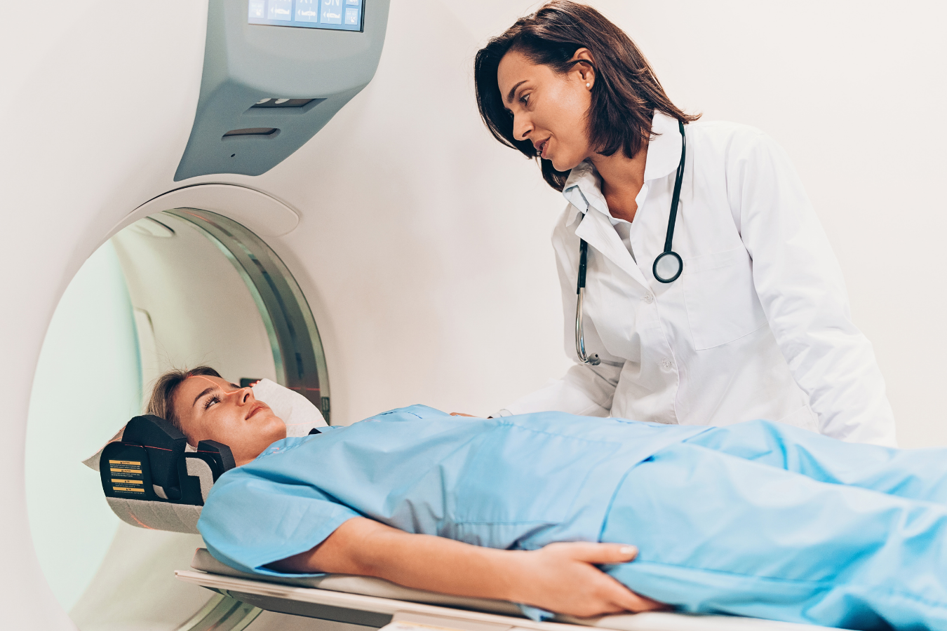 Female patient lying on an MRI table, about to be put inside the MRI tube. Female healthcare provider with a stethoscope around her neck, standing above the patient.