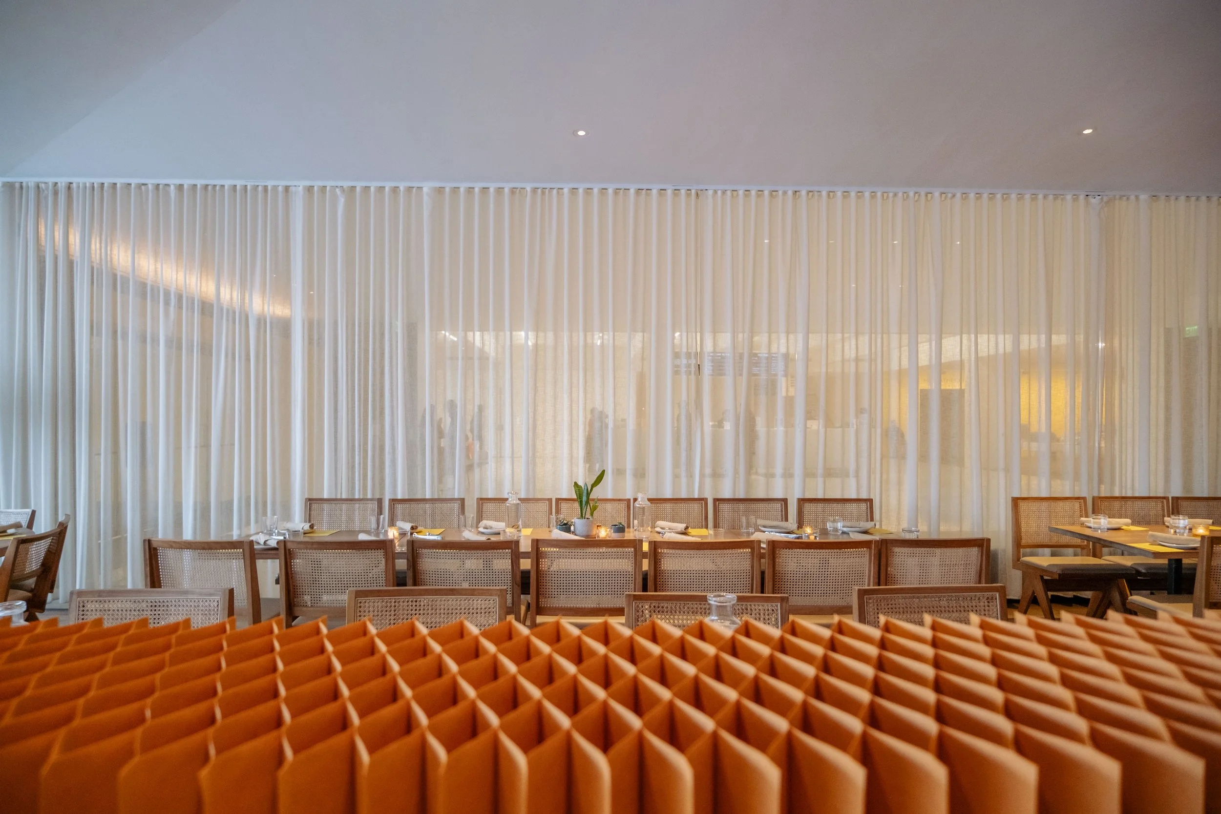 Empty restaurant dining area with a long table set with glasses, plates, and a small plant in the center, behind a decorative orange partition and separated by white sheer curtains.
