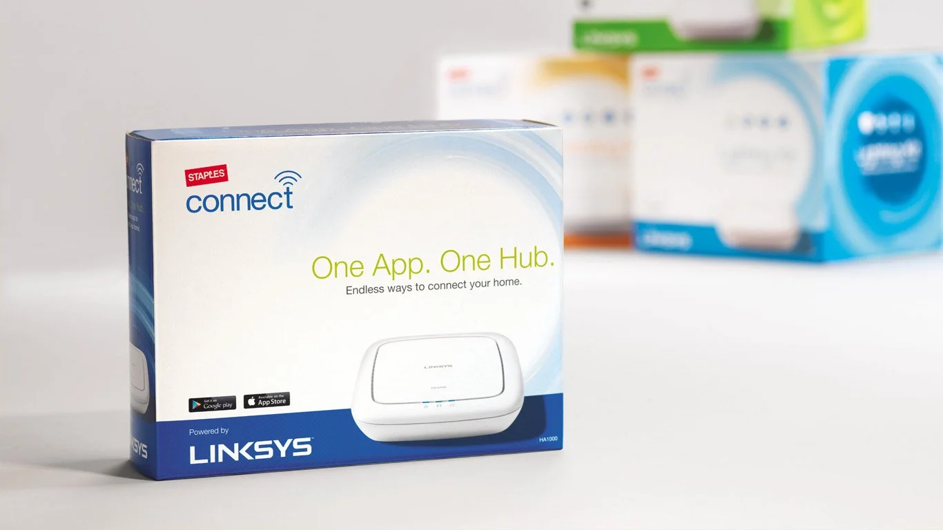 Package design for Staples Connect smart home hubs with tagline 'One App. One Hub.' Blurred boxes in the background.