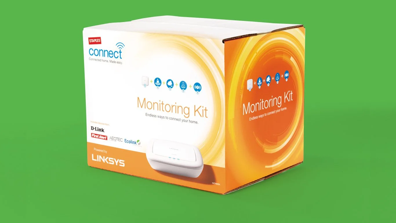 A boxed Staples Connect Monitoring Kit on a green background, featuring smart home integration devices from brands like D-Link, First Alert, Aeotec, and Ecolink, and powered by Linksys.