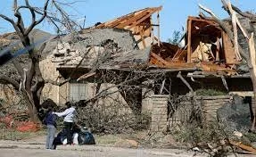 A large tree has fallen onto a damaged house, causing significant destruction. Three people are nearby, observing the aftermath, with a clear blue sky in the background.