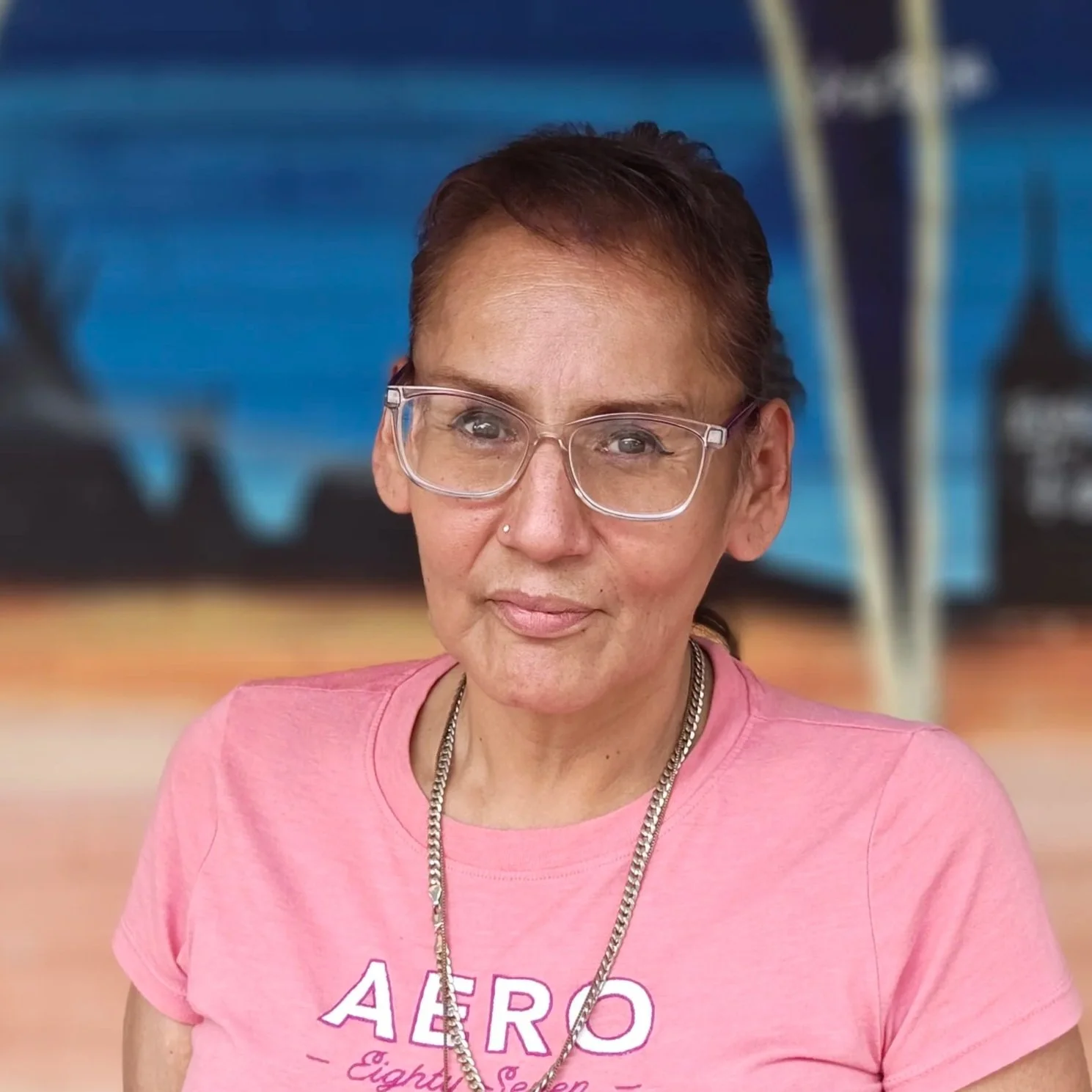 Woman wearing glasses and a purple shirt with a smile, standing in front of a colorful mural.