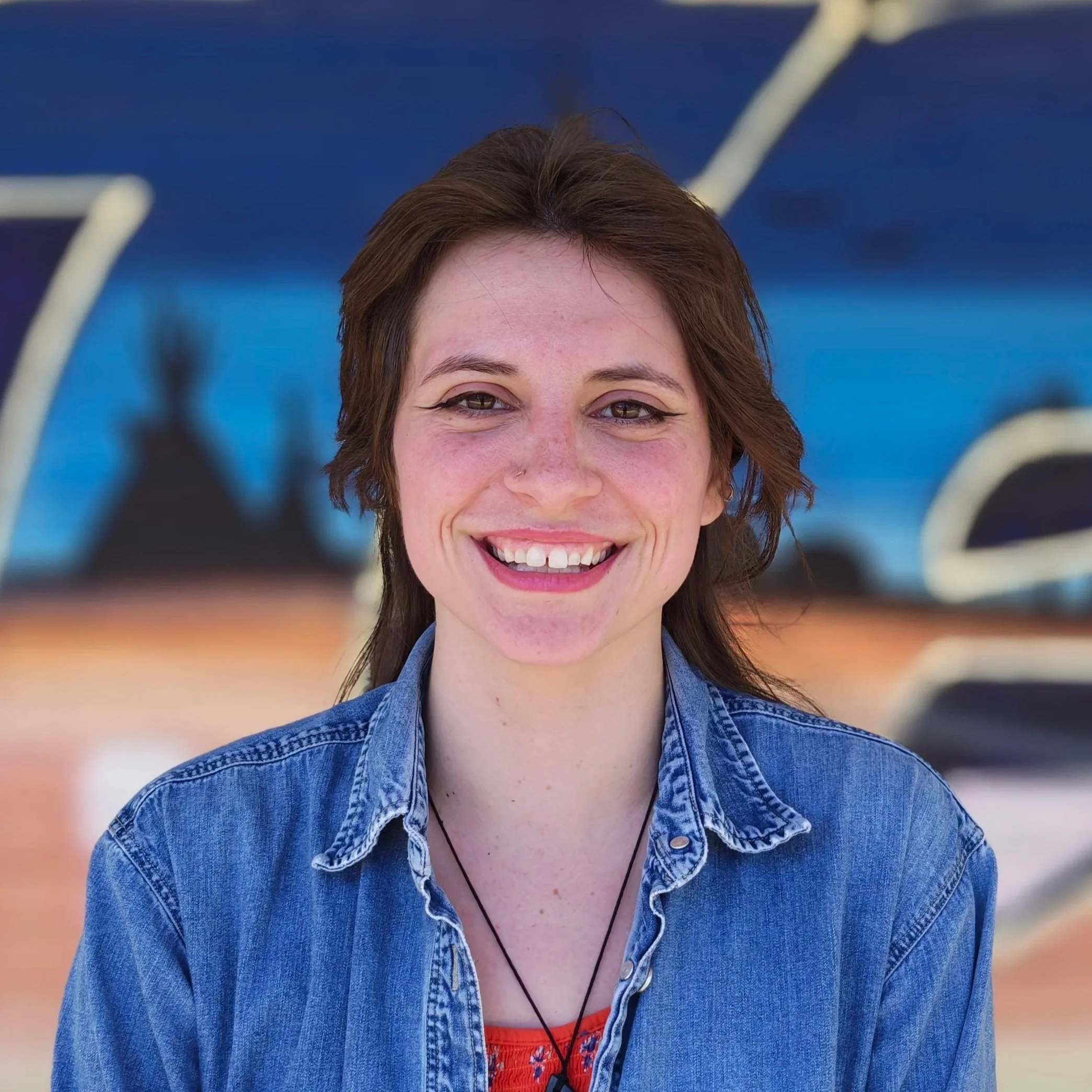 A smiling woman with short brown hair wearing a denim shirt and red top, standing outdoors with blurred background.