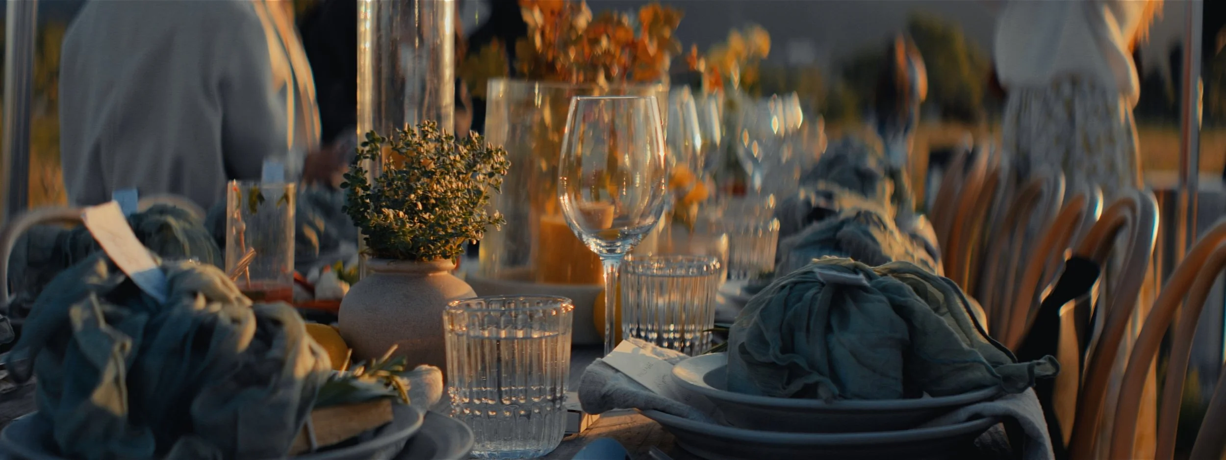 A close-up of a dining table set outdoors during sunset, with glassware, plates, cloth napkins, a potted plant, and floral centerpieces, with blurred people in the background.