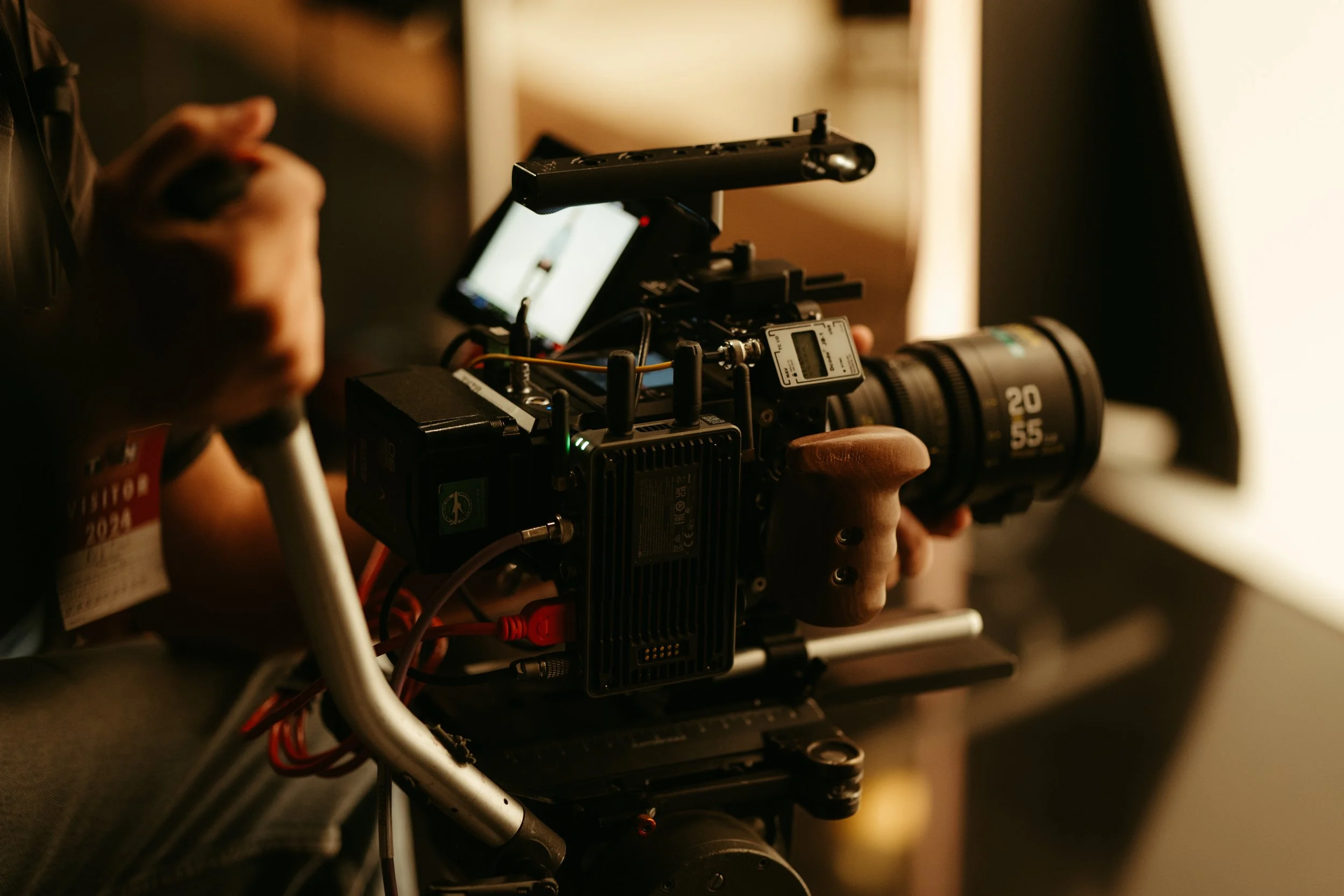Close-up of a professional video camera with a large lens and monitor, being operated by a person in a dimly lit setting.