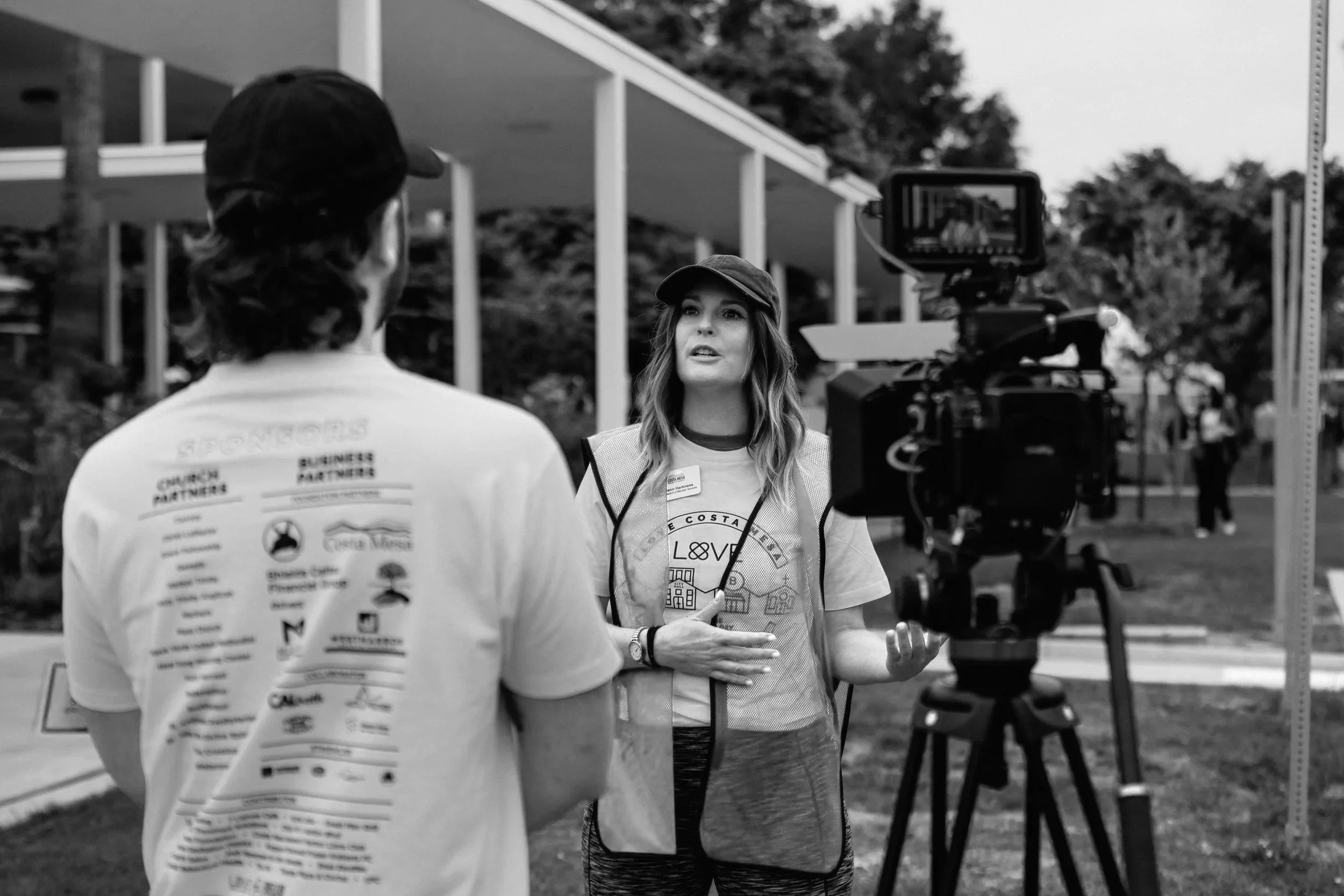 A woman being filmed during an interview outdoors with a video camera, with a man speaking to her, both wearing casual clothing.