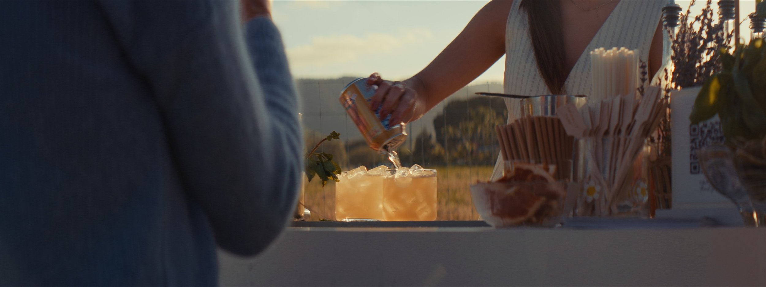 Person pouring a beverage into a clear glass on an outdoor table at sunset, with snacks and utensils nearby.