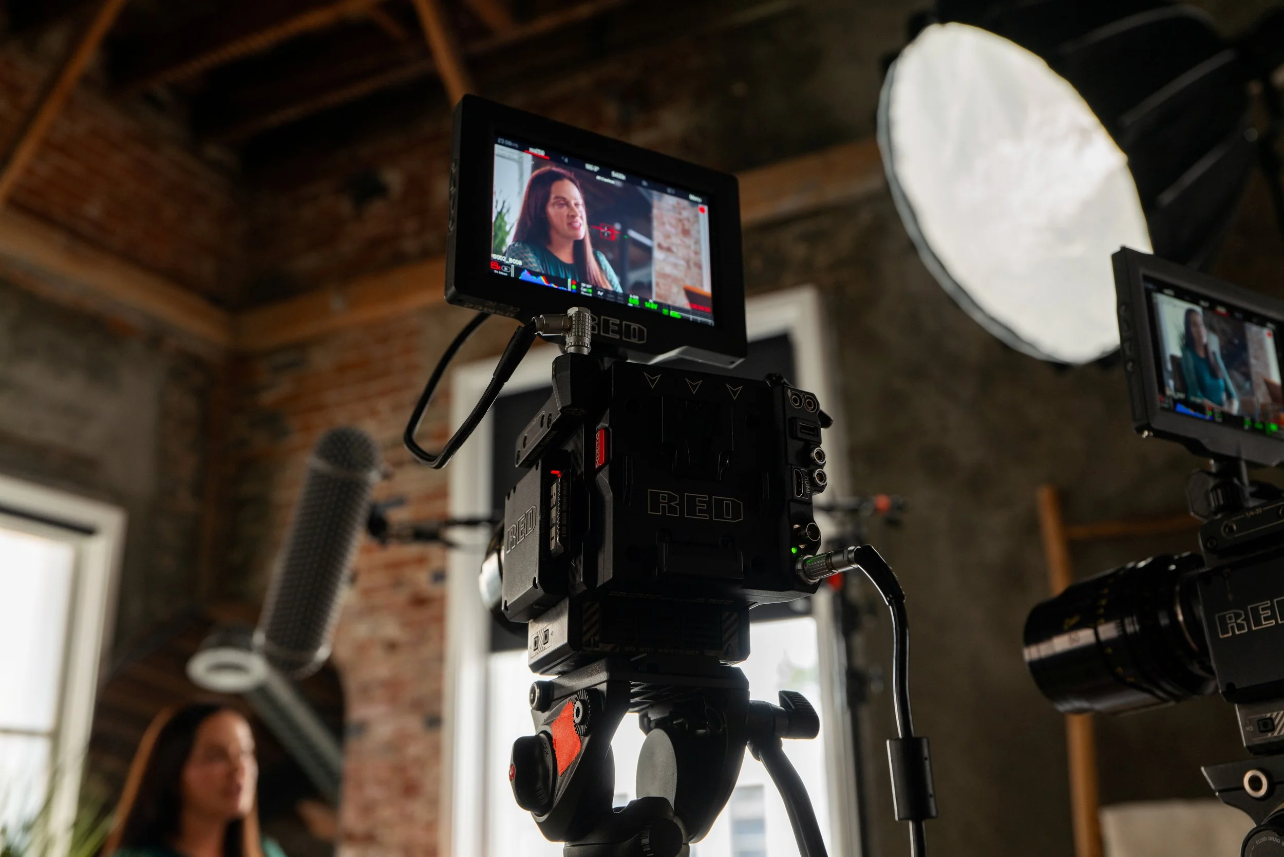 Video camera filming a woman during an interview or recording session in a room with brick walls and large windows.