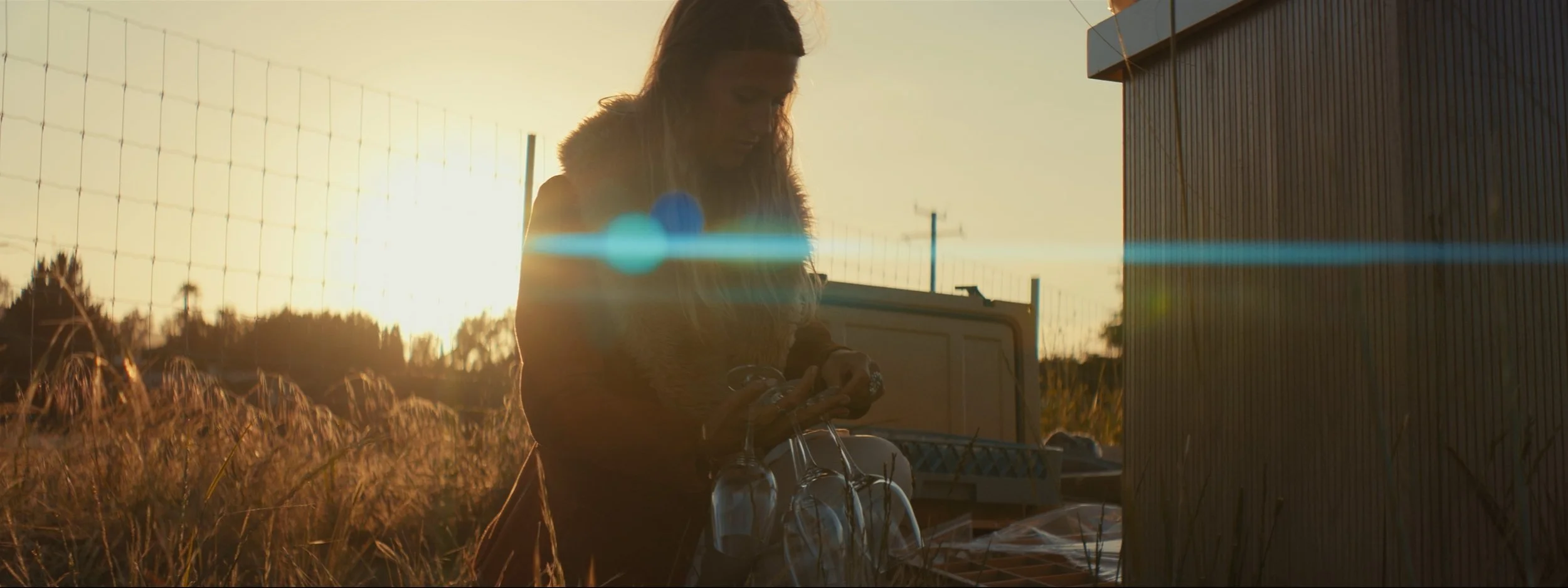 A woman with long hair standing outdoors during sunset, looking down at her phone, with a basket of jars and a bicycle nearby, surrounded by tall grass and a metal building.