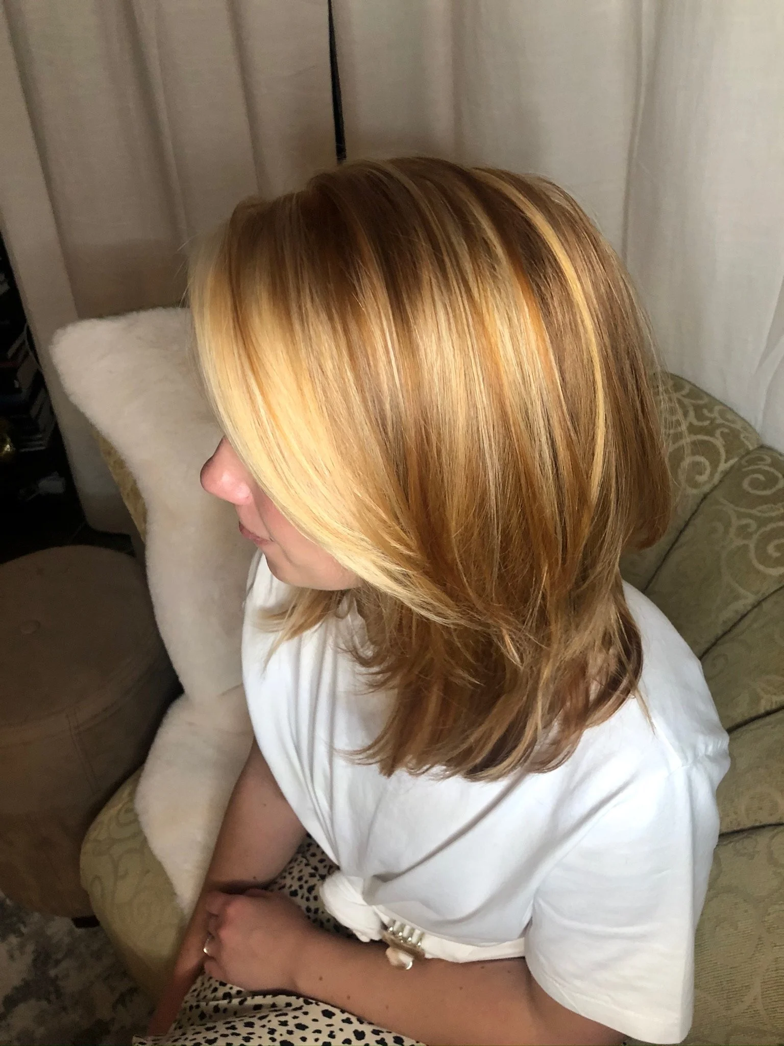 A woman with shoulder-length highlighted blonde hair sitting on a beige patterned couch, wearing a white shirt.