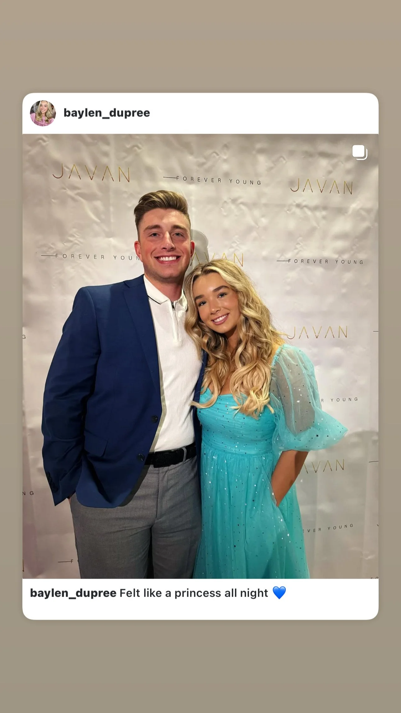 A young man and woman smiling and posing together at an event with a Javan backdrop. The man is wearing a navy blazer and gray pants, and the woman is in a turquoise princess gown with long, wavy blonde hair.
