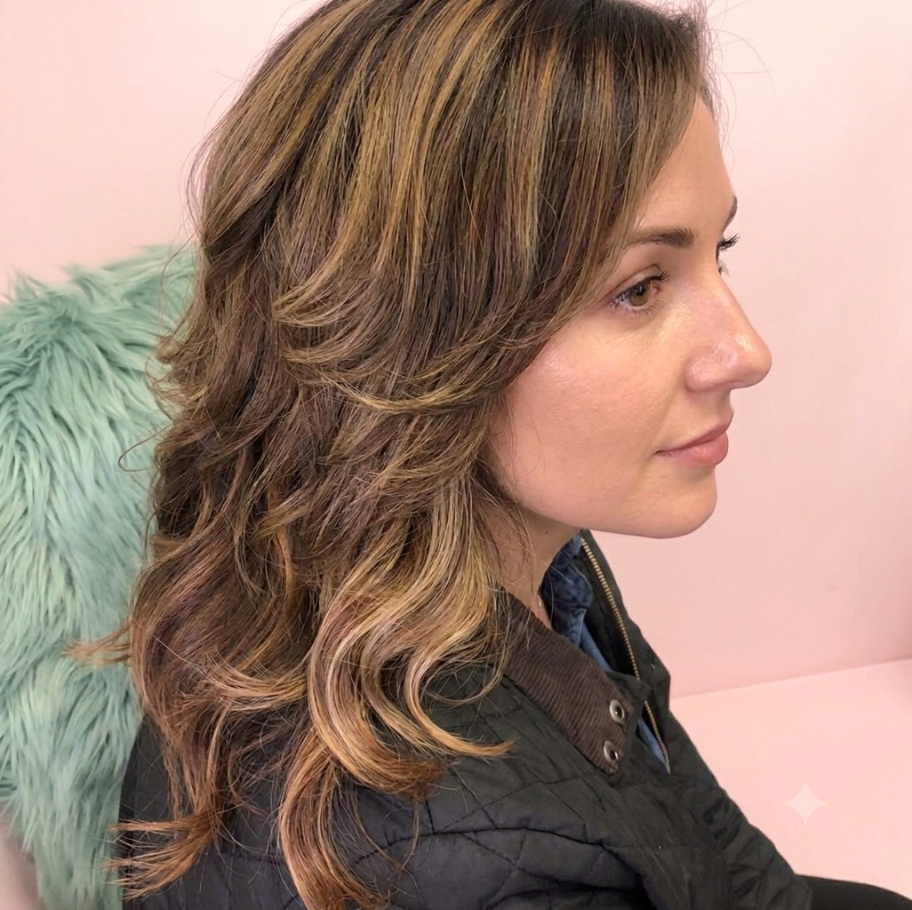 Profile of a young woman with wavy light brown hair and subtle highlights, sitting on a pastel pink background with a light blue furry cushion nearby.