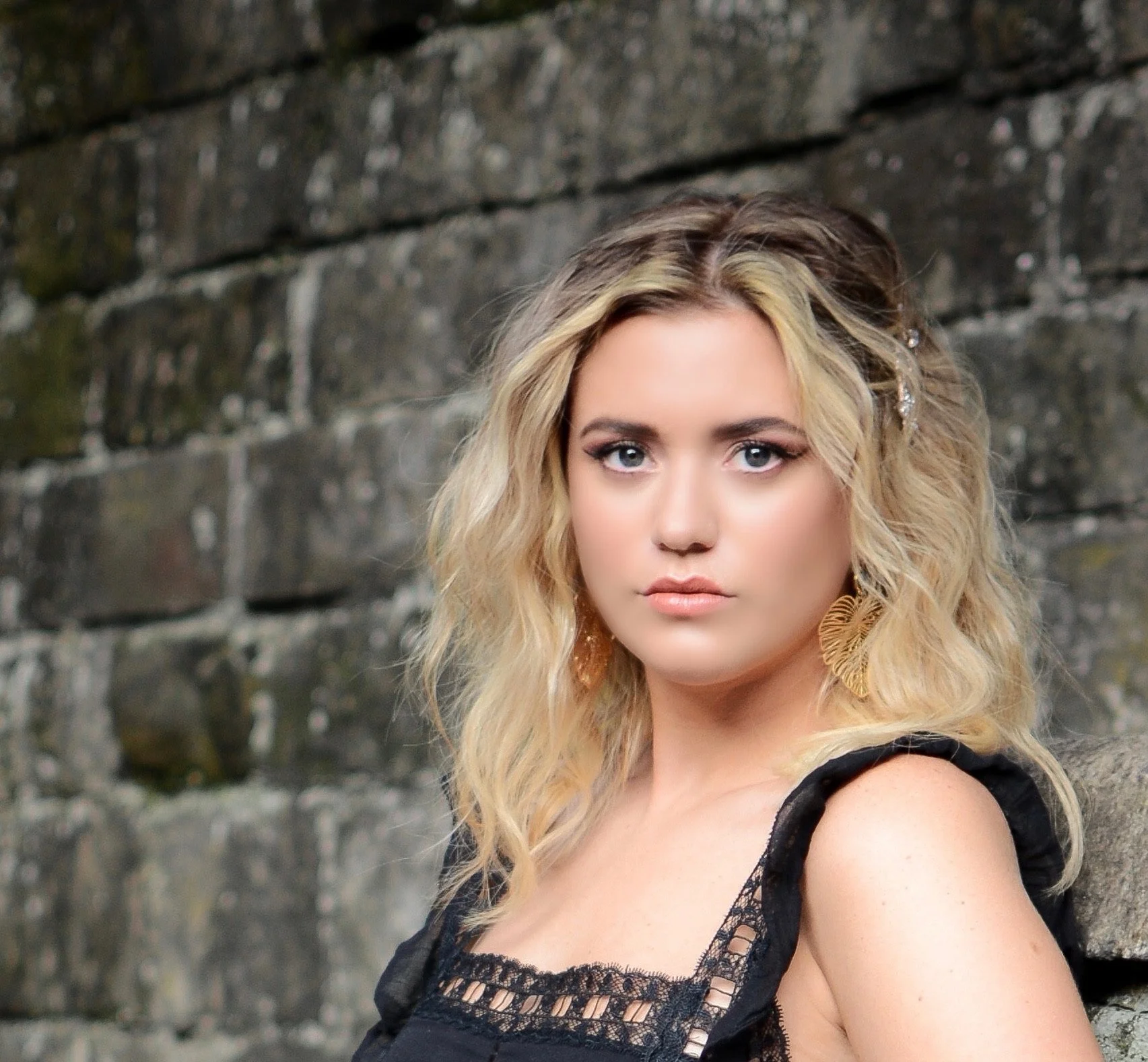 A woman with blonde, wavy hair and blue eyes standing in front of a stone wall, wearing a black lace top and large feather-shaped earrings.