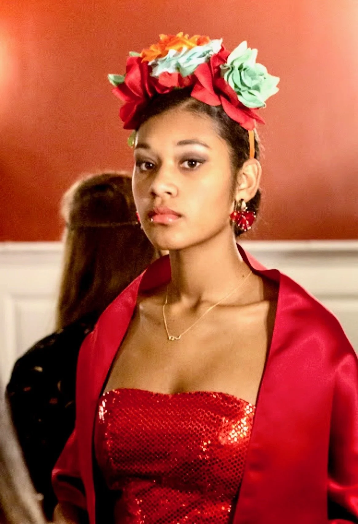 A young woman with short dark hair wearing a red sequined top, red satin jacket, a flower crown with red, green, and orange flowers, and red earrings, standing in front of a red wall.