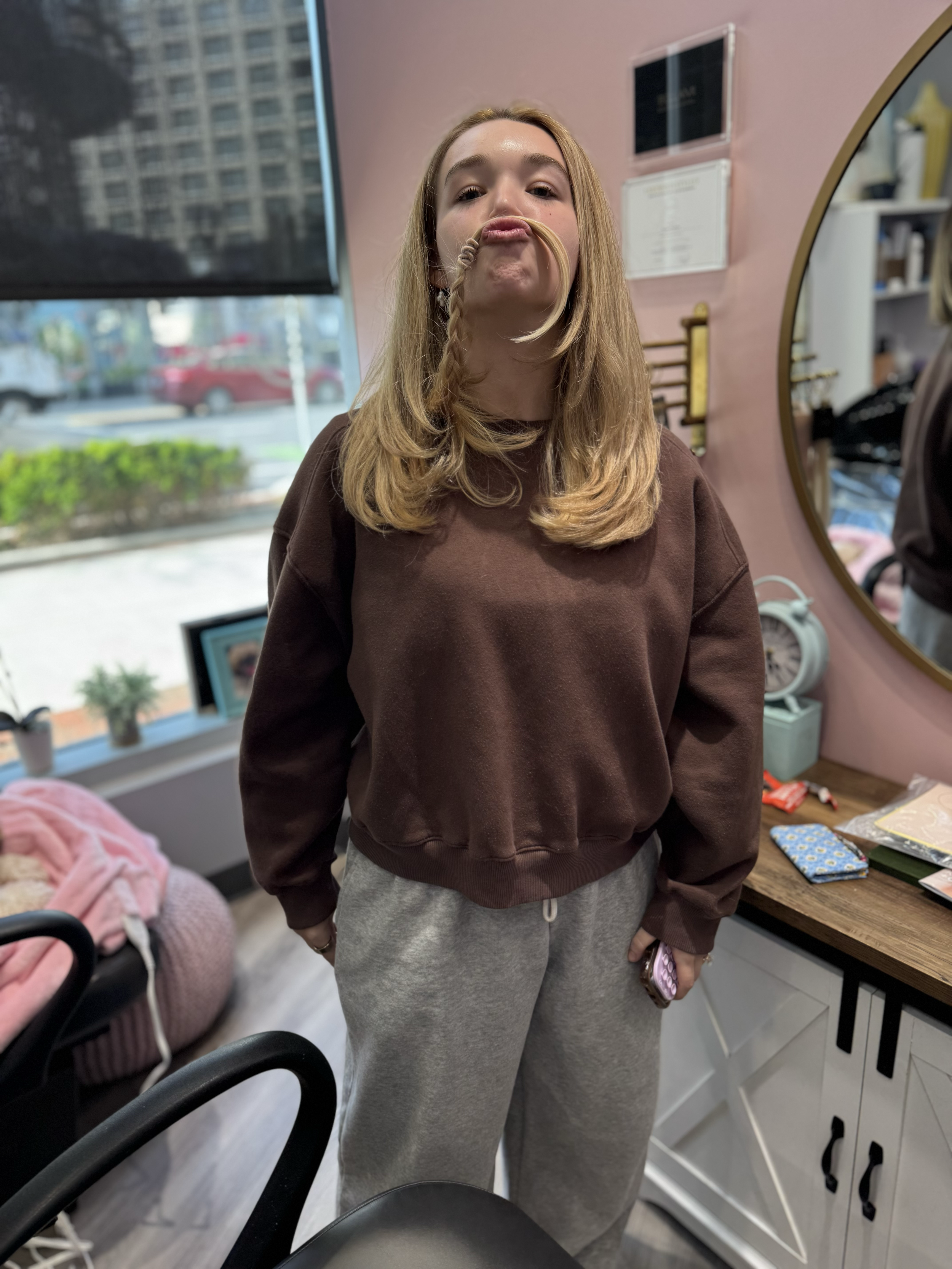 Baylen Dupree-Dooley with long blonde hair making a duck face for the camera, wearing a brown sweatshirt and gray sweatpants, in a room with pink walls and various objects, including a large mirror, a window, and a table with items on it.