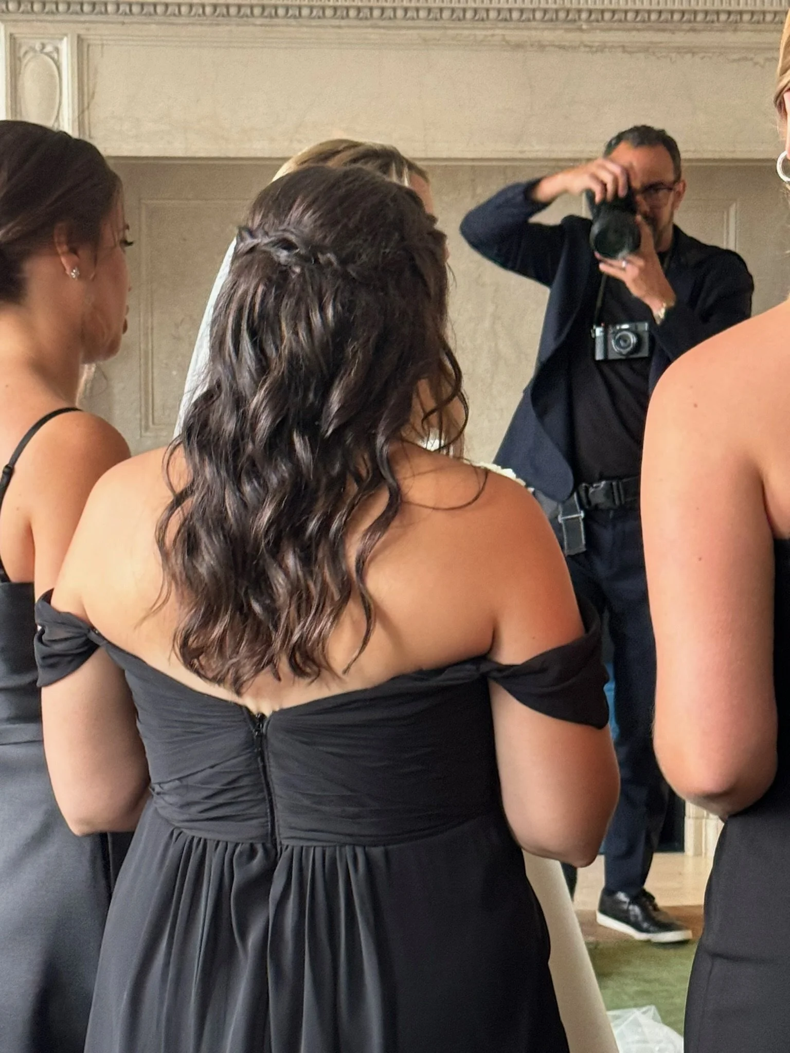 People at a formal event, including women in black dresses and men in dark suits, with a woman in a black dress prominently in the foreground and a photographer taking pictures.