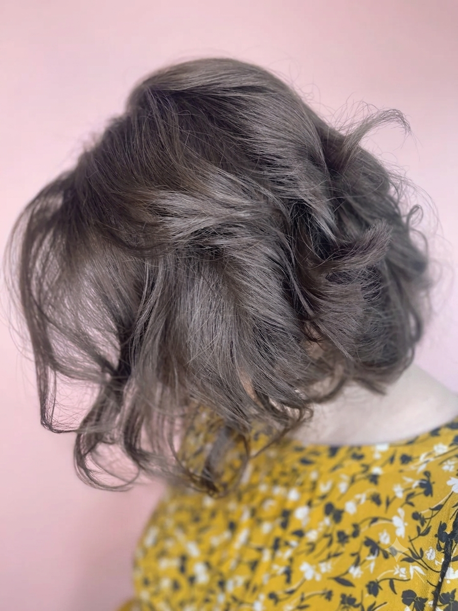 Close-up of a person with short, wavy gray hair wearing a yellow top with a floral pattern, against a pink background.