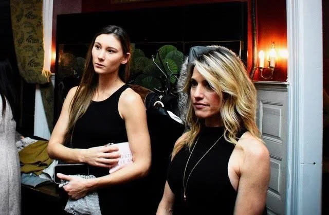 Two women with blonde and brunette hair standing indoors, one holding a piece of paper, in a room with red walls and white wainscoting, lit by warm lamps.
