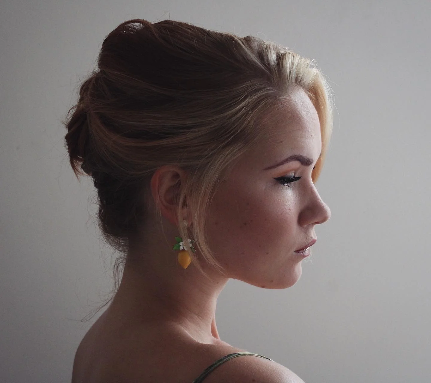 Profile of a young woman with braided blonde hair, eye makeup, and strawberry earrings, standing against a plain wall.