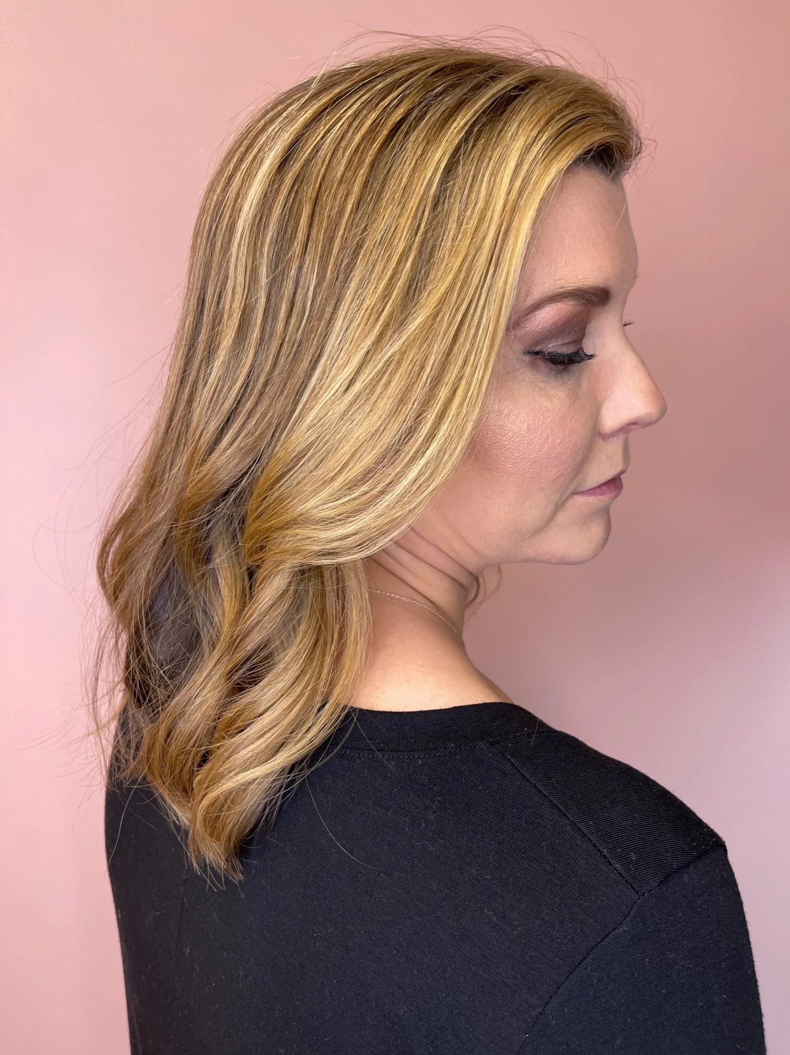 Close-up side profile of a woman with long, blonde, wavy hair wearing a black top and standing against a pink background.