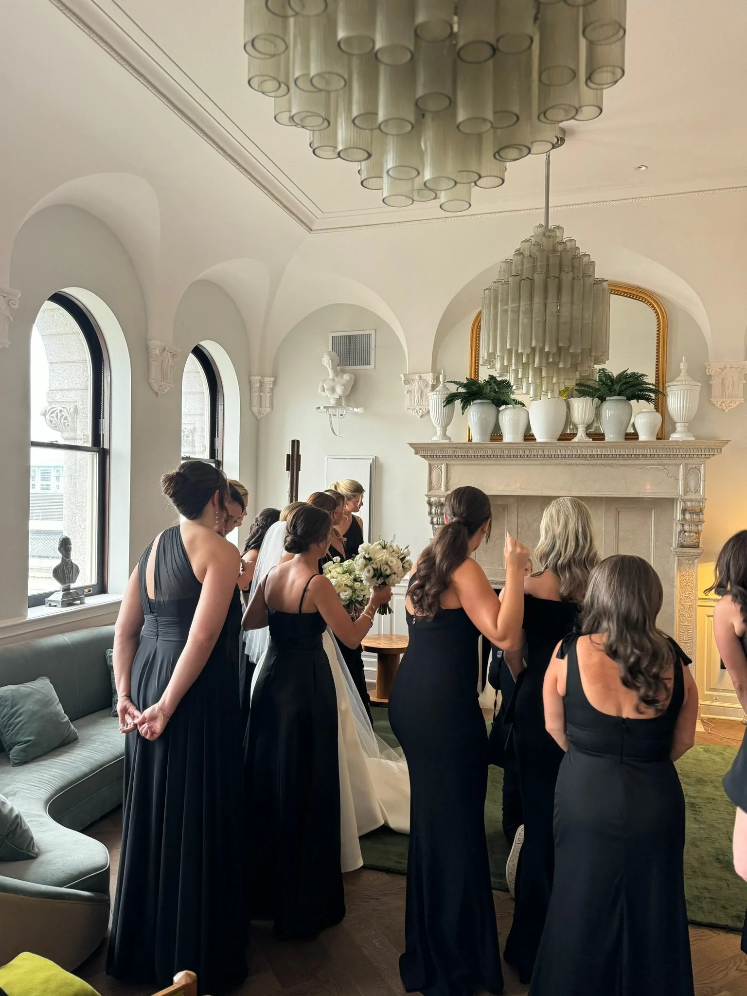 A group of women dressed in black evening gowns gathered indoors, some holding bouquets, in an elegant room with arched windows, a fireplace, and decorative elements.