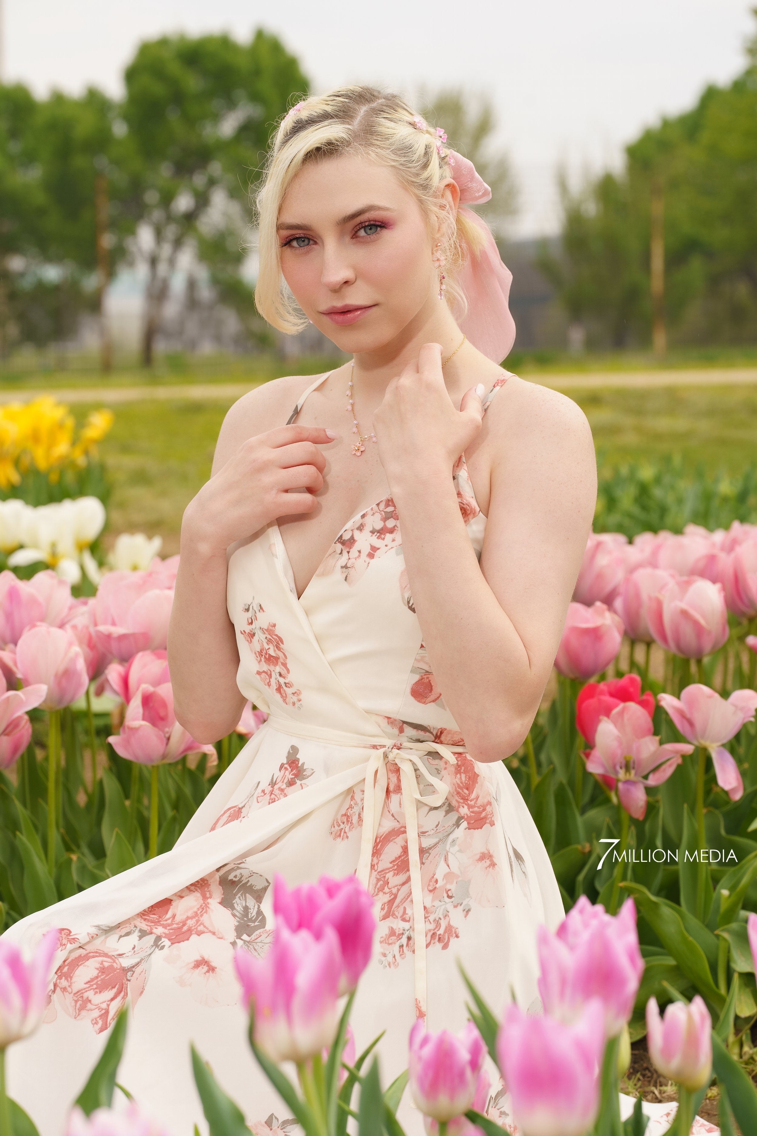 A young woman with blonde hair and pink makeup standing among pink tulips in a garden on a cloudy day. She is wearing a sleeveless, floral-patterned dress and jewelry, and looking at the camera with a calm expression.