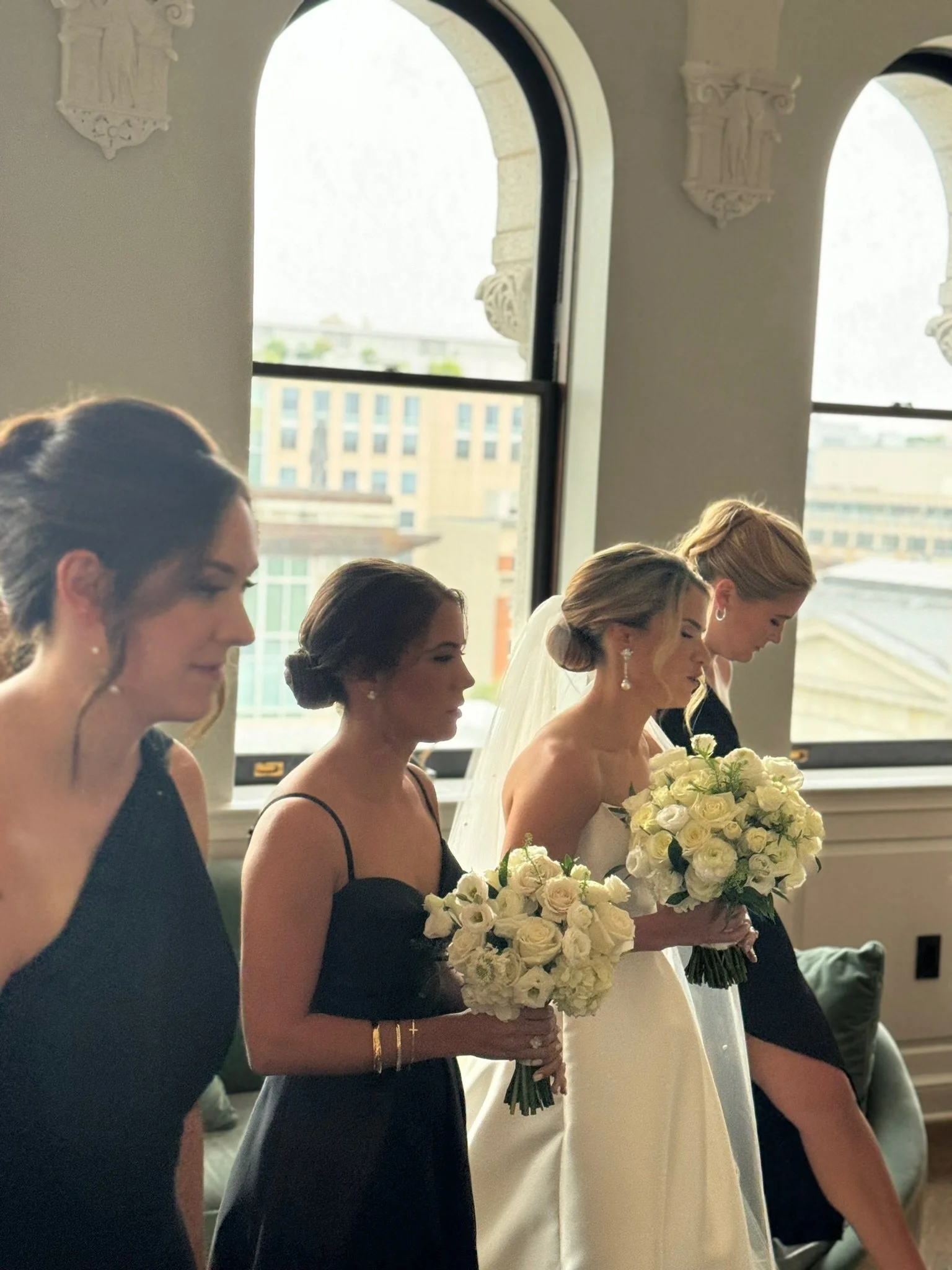 Four women, including a bride in a white wedding dress with a veil, standing in a row holding bouquets of white and cream flowers during a wedding ceremony inside a room with large windows and decorative architectural elements.
