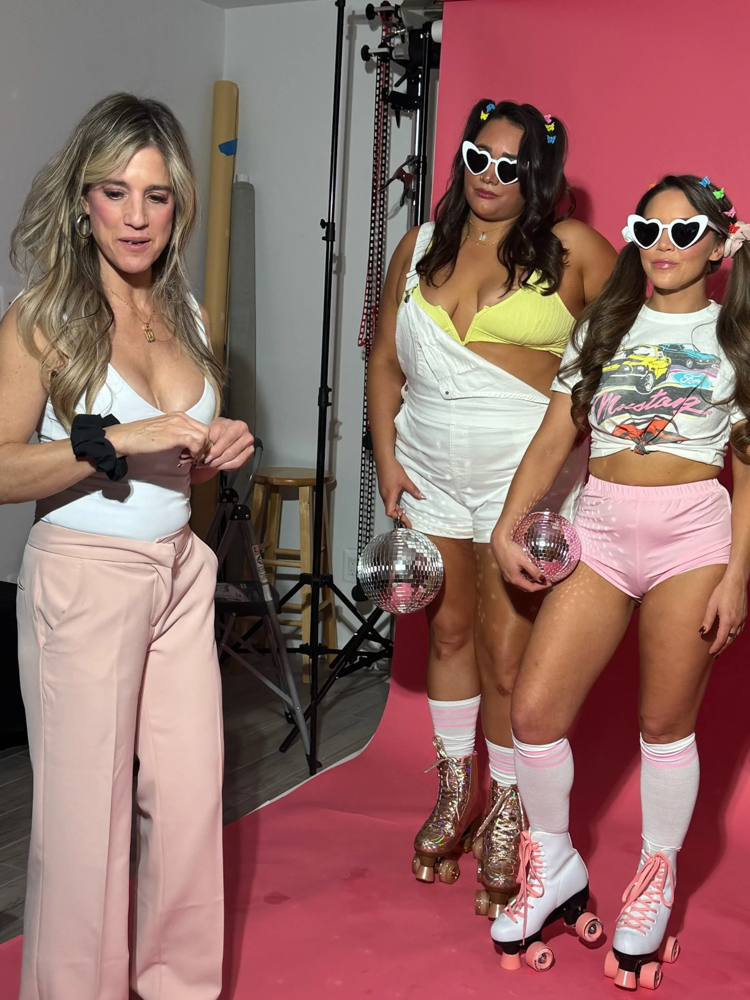 Three women are in a photo shoot, with pink background. The woman on the left has blonde hair, wearing a white tank top and pink high-waisted pants. The two women on the right have long brown hair, are dressed in colorful roller skating outfits with 