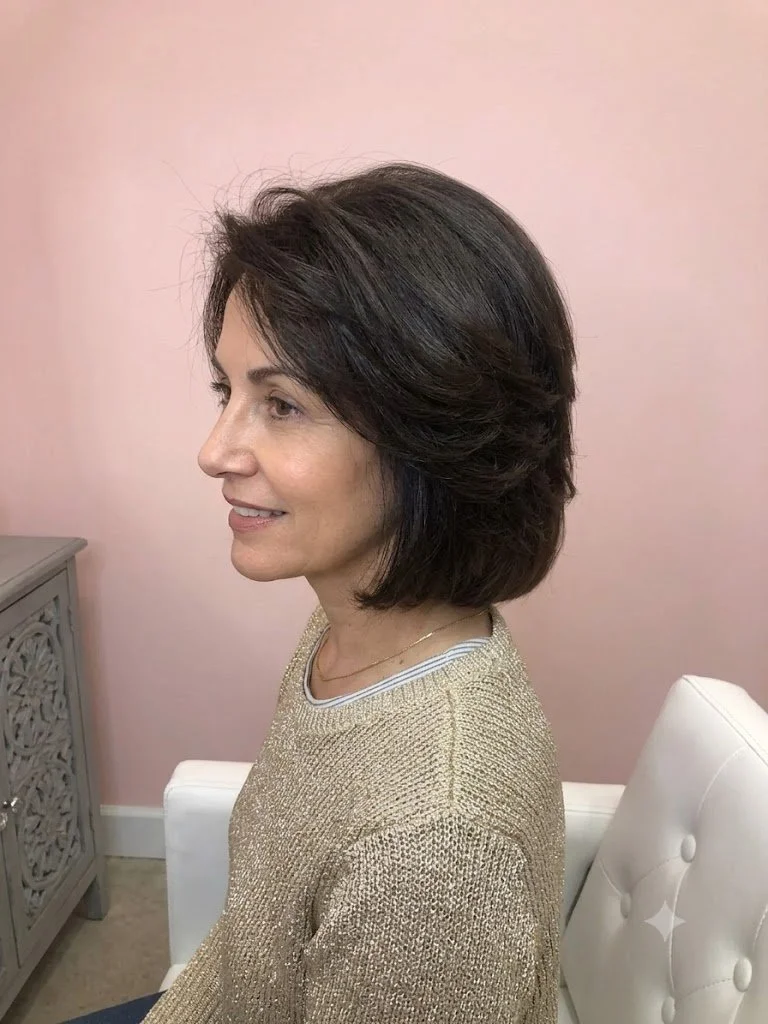 Profile of a woman with dark brown, layered bob hairstyle, wearing a gold sweater, sitting in a room with a pink wall and white furniture.