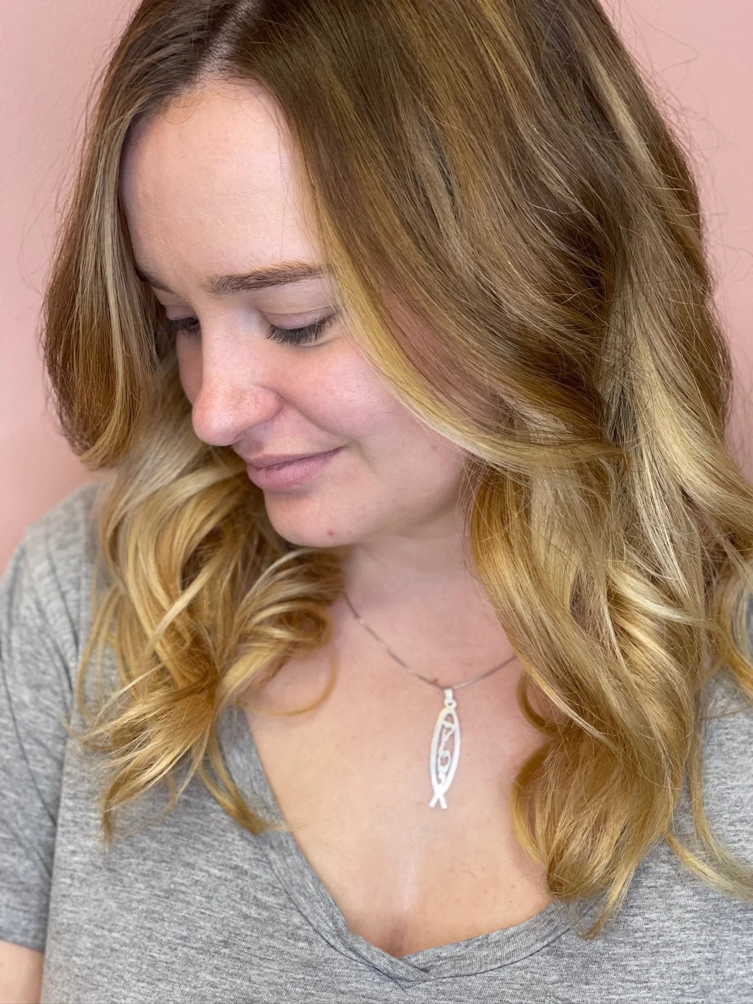 A woman with long, wavy blonde hair looking down, wearing a gray top and a necklace with a pendant, standing against a pink background.