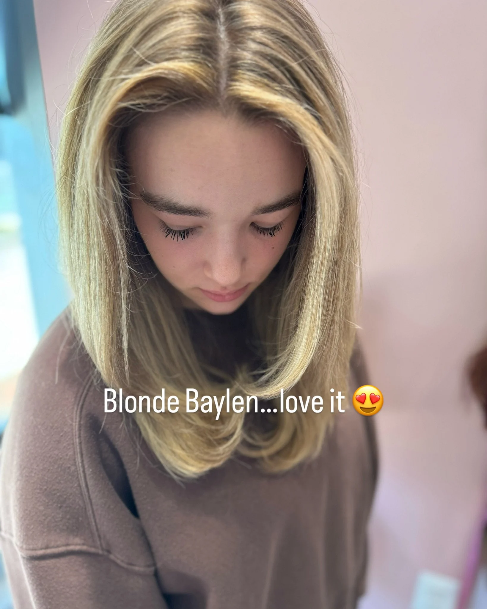 A woman with blonde hair styled in loose waves, looking down, wearing a taupe-colored top or dress.