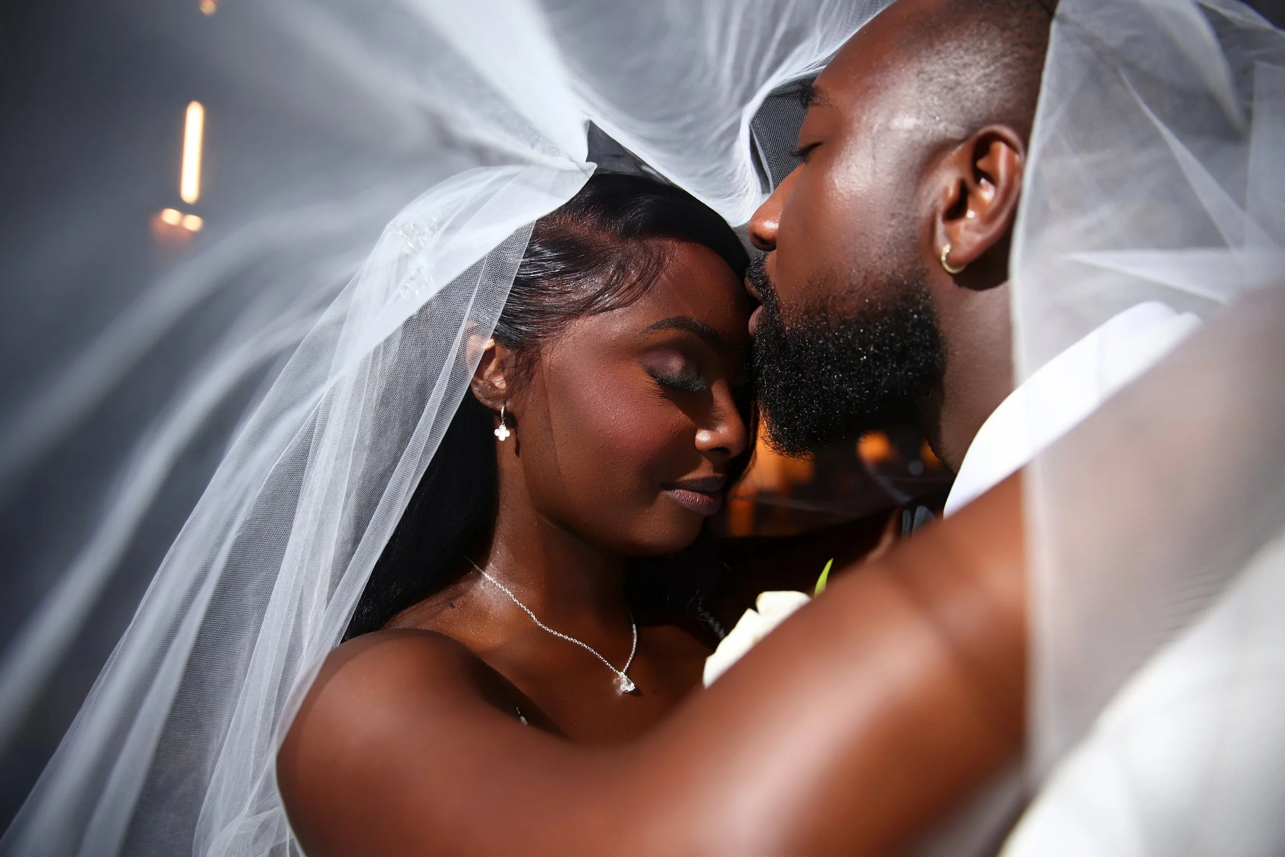 A close-up of a newlywed couple with eyes closed, nestled under a veil, sharing an intimate moment. The bride has dark skin, black hair, and is wearing earrings and a necklace. The groom, also with dark skin, has a beard, earrings, and is dressed in 