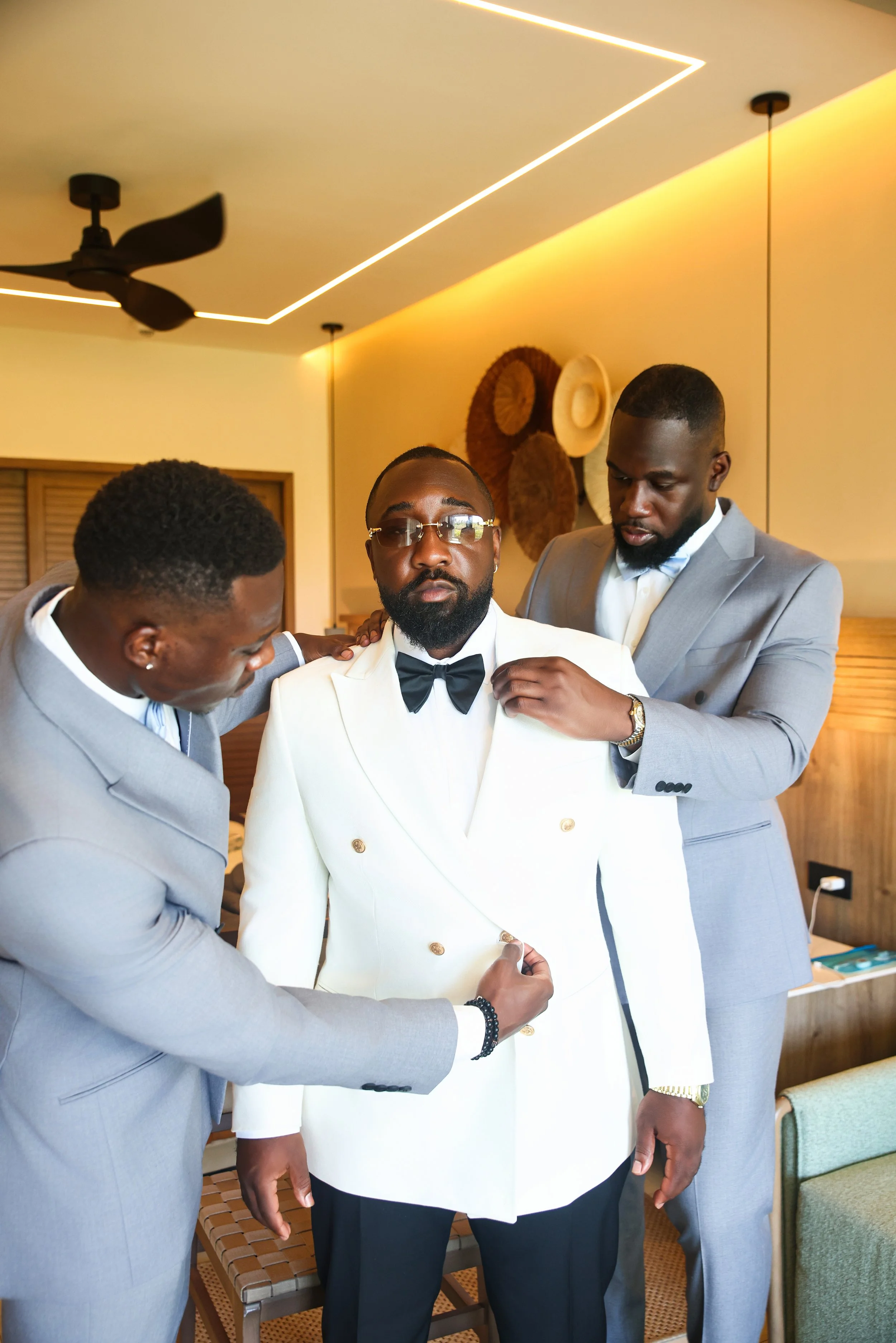 Three men in formal attire, one adjusting another's tuxedo, in a warmly lit room with decorative hats on the wall.