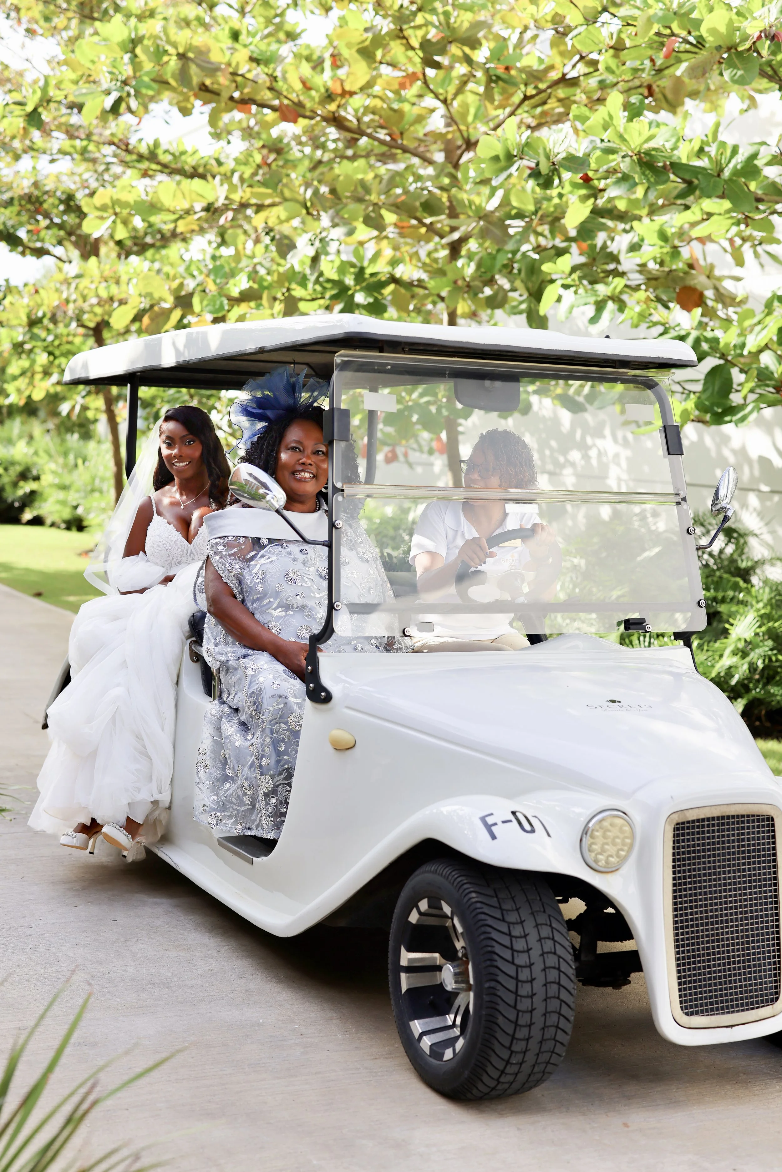 Women in wedding dress and traditional attire riding in a golf cart outdoors near green trees and grass.