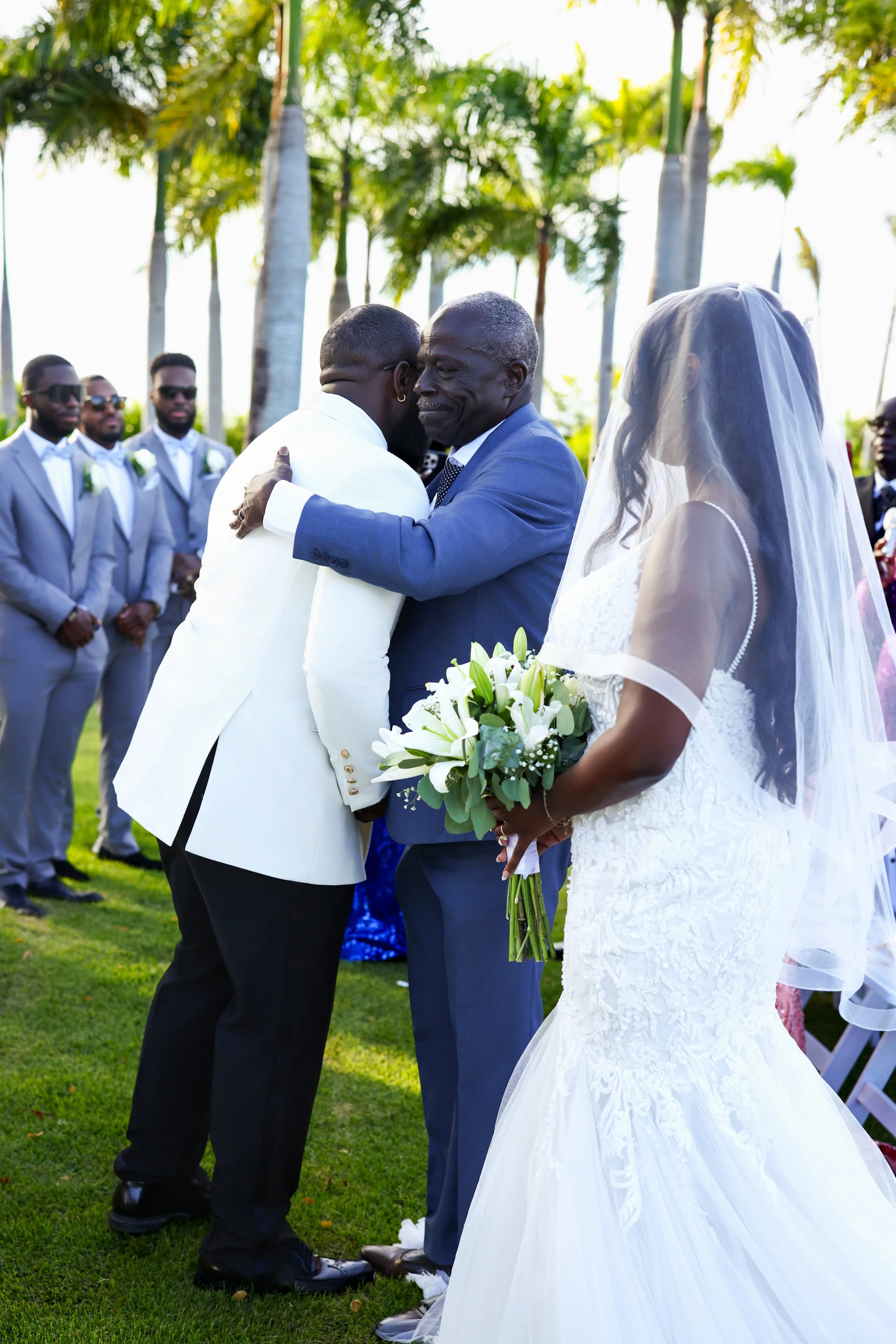A wedding scene outdoors with a groom in a white tuxedo hugging an older man in a blue suit, a bride in a white lace wedding gown and veil holding a bouquet of white lilies, and groomsmen in light gray suits with white shirts and black sunglasses sta