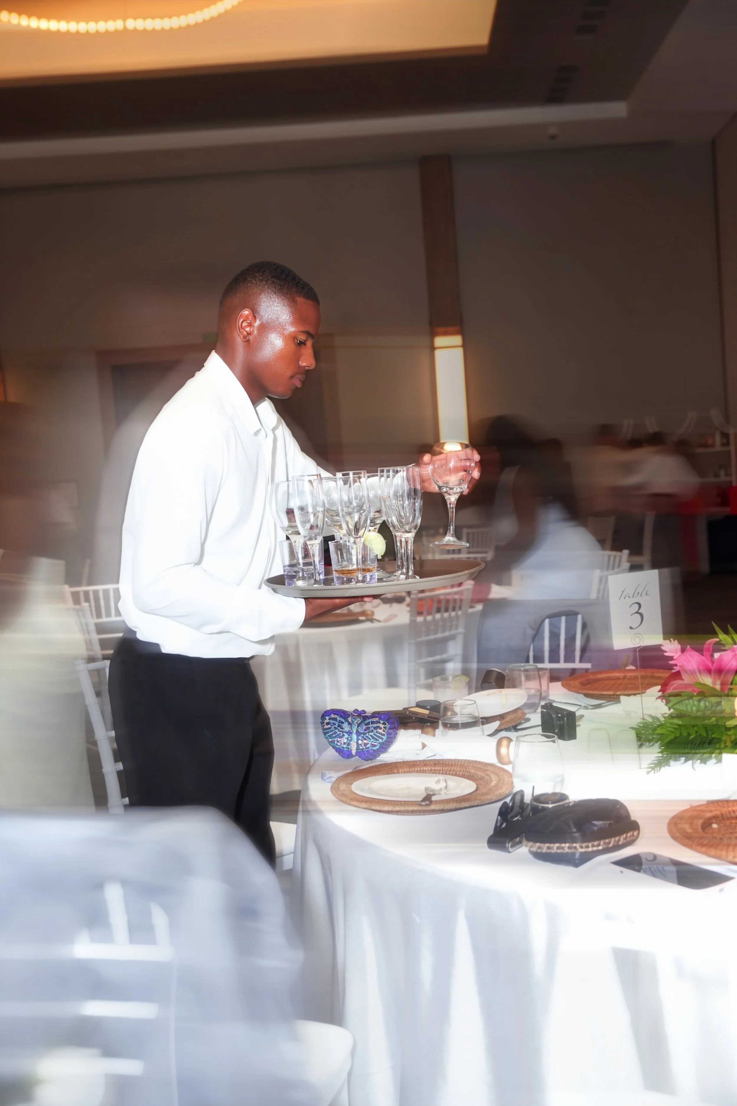 A young man in a white shirt and black pants serving drinks at a formal event, with a tray of glasses and a champagne glass in his hand, in a brightly lit room with white chairs and decorated tables.